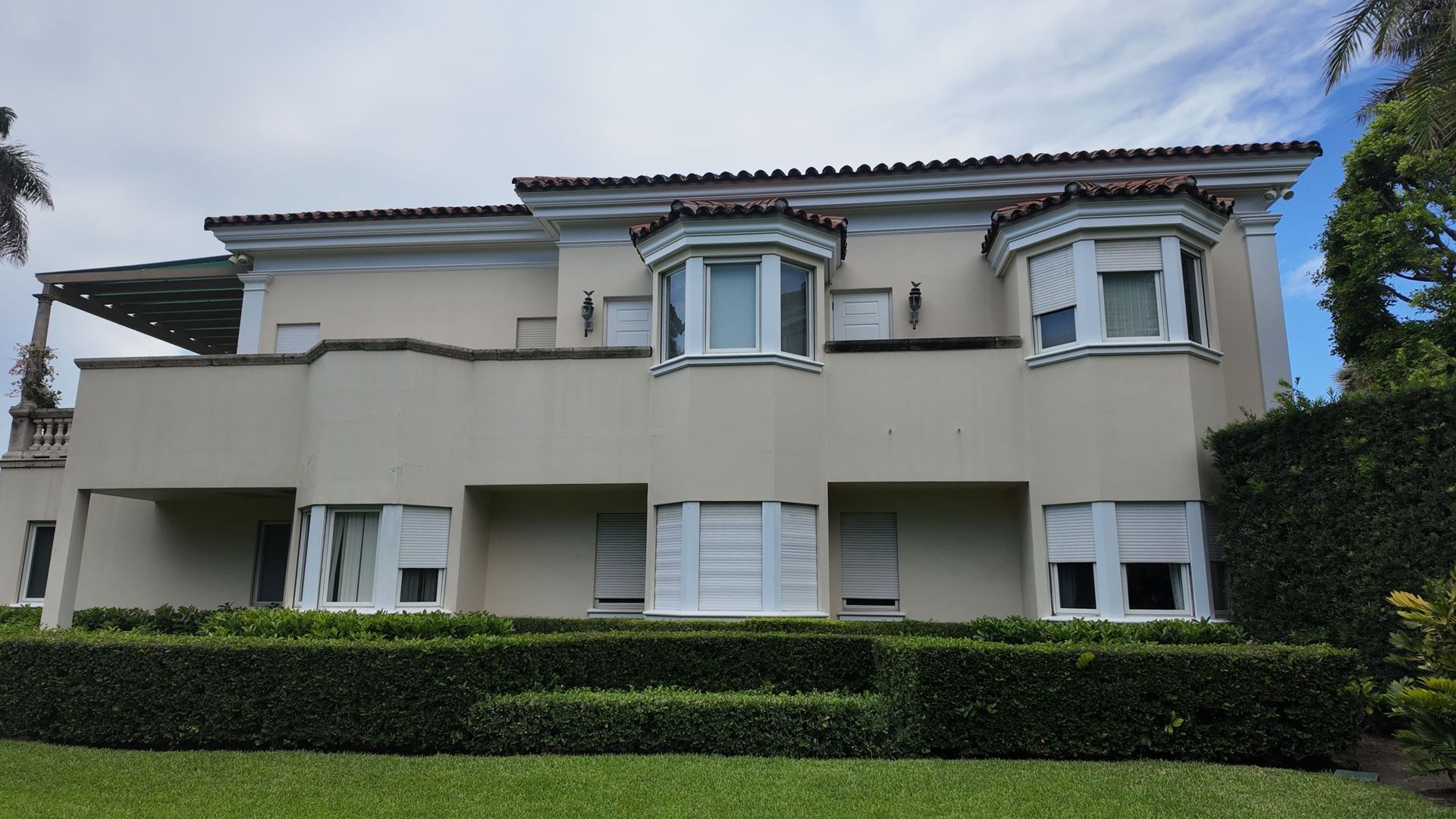 Two-story beige house with white trim, front hedge, and a cloudy sky overhead