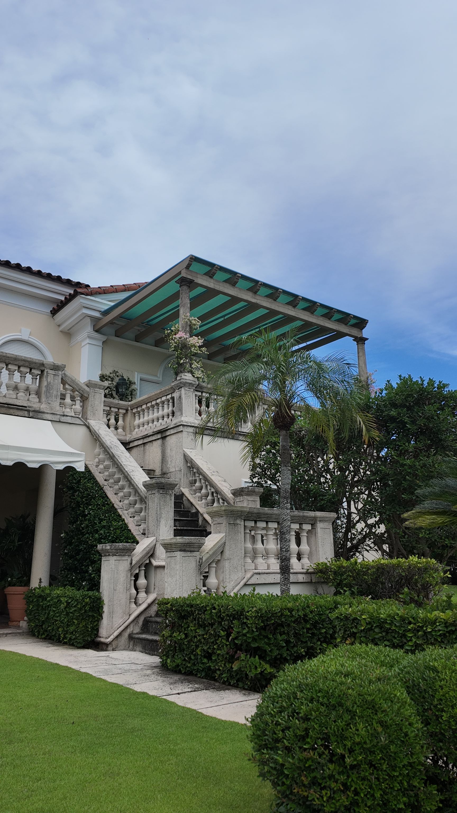 White villa with outdoor staircase, green awning, and manicured hedges under a blue sky.