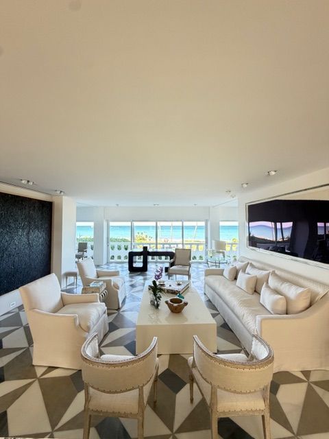Modern white living room with sofas, chairs, geometric rug, ocean-view windows, and a large TV