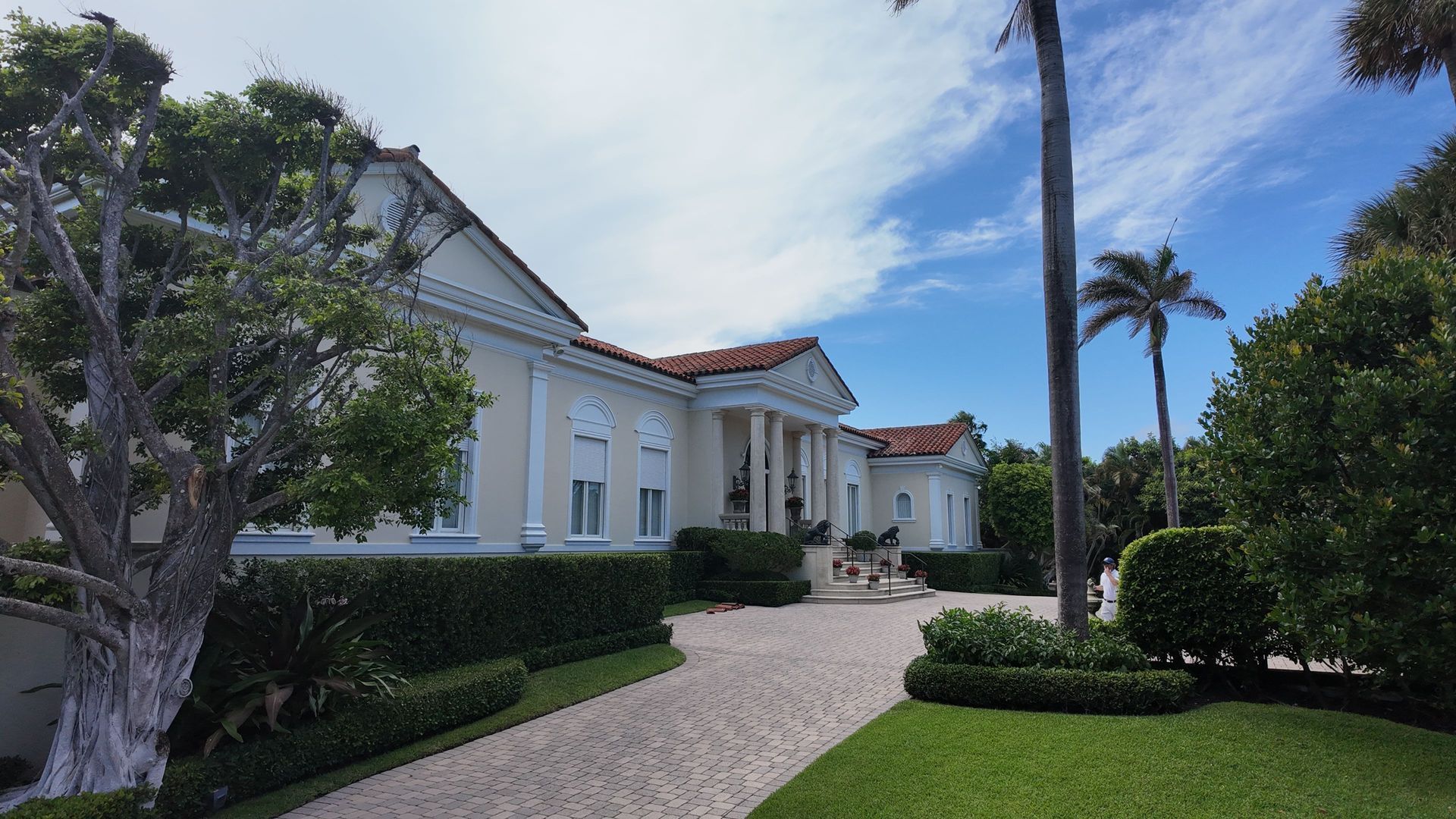 White Mediterranean-style house with red-tiled roof, palm trees, and a curved gravel driveway under a blue sky