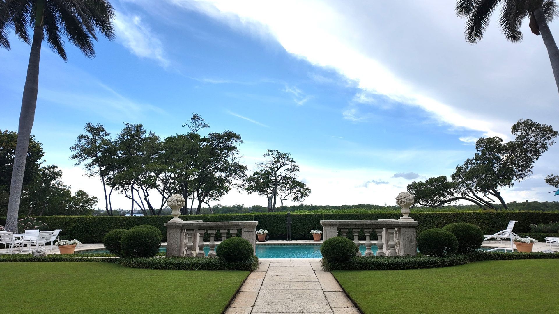 Garden terrace with a stone walkway, trimmed hedges, fountains, and trees under a blue sky
