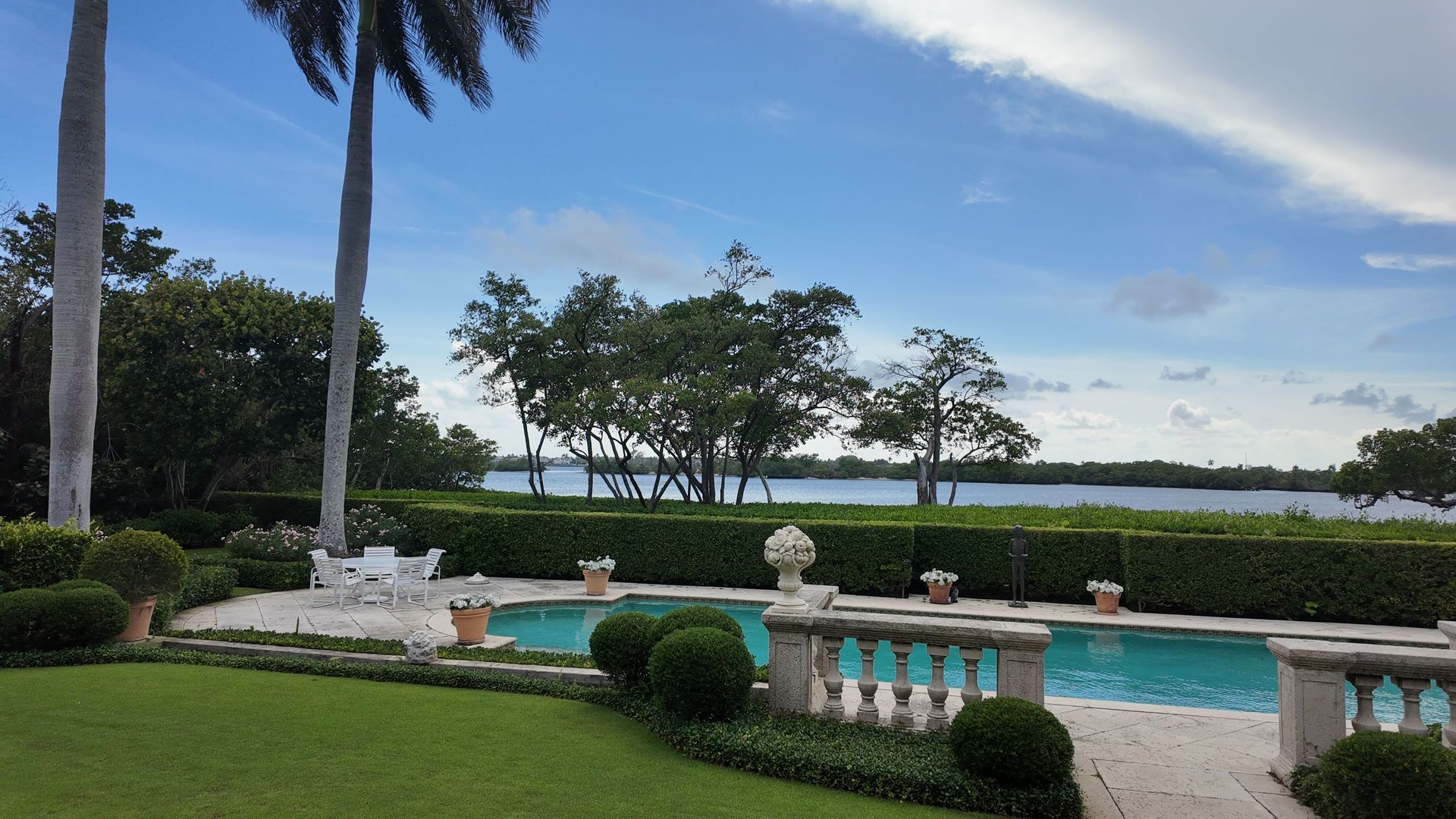 Poolside patio overlooking a calm waterfront, with white balustrades, palm trees, and trimmed hedges.