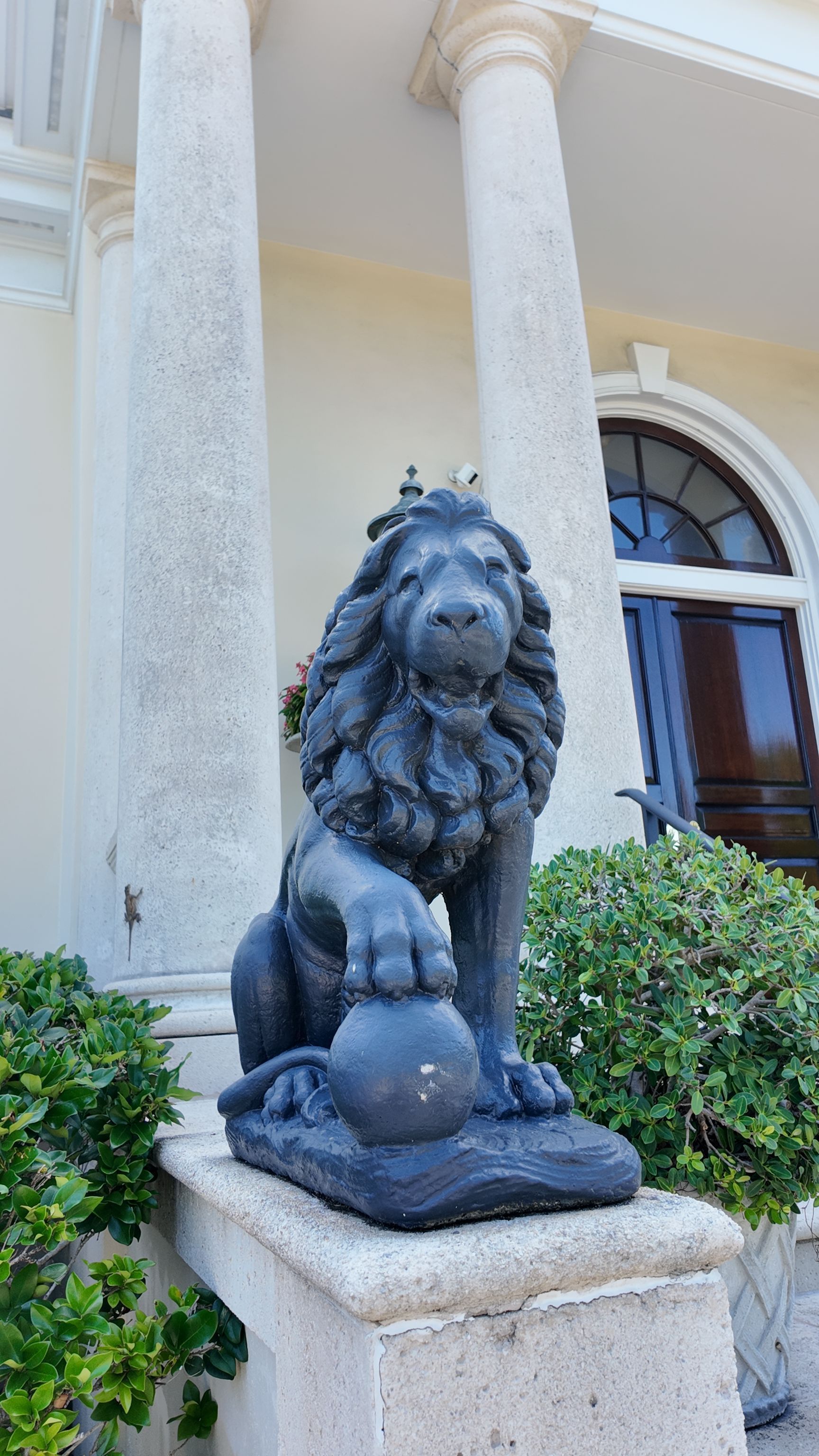 Stone lion statue on a pedestal outside a building with cream columns and an arched window.