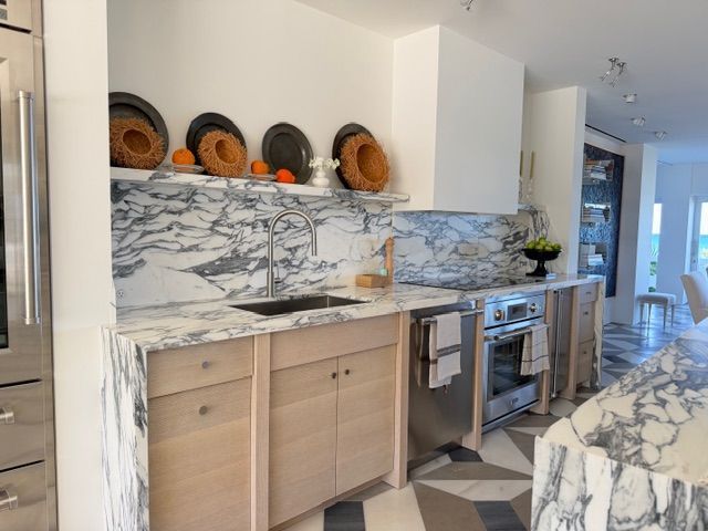 Modern kitchen with marble countertops, light wood cabinets, and woven decor on open shelves.