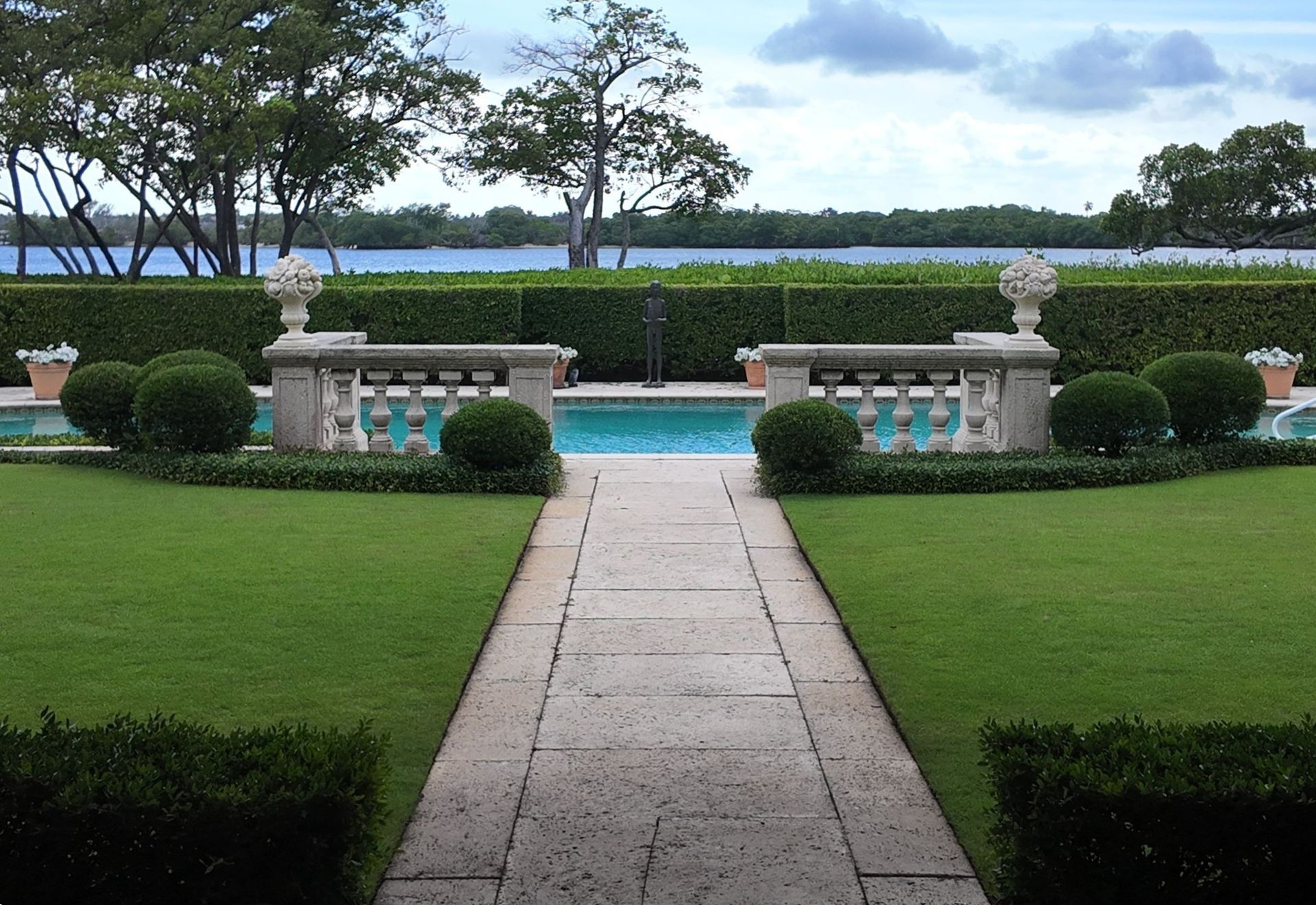 Stone walkway through manicured lawn toward a pool and hedge, with benches and trees beyond.
