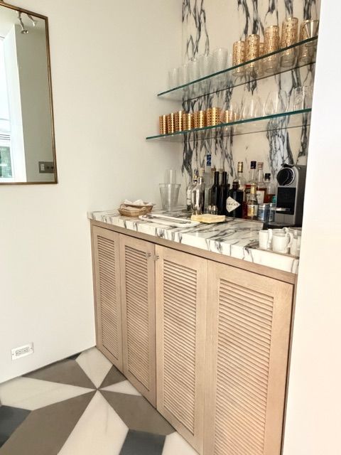 Modern bar cabinet with bottles, glass shelves, and marble top in a bright room with geometric tile floor