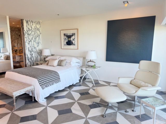 Modern bedroom with white bed, lounge chair, geometric rug, and large dark wall art