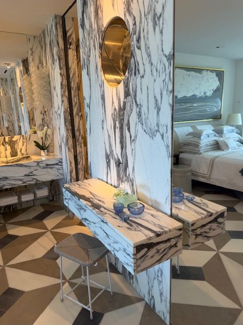 Marble vanity area with gold wall mirror, geometric floor tiles, and a bed reflected in the mirror