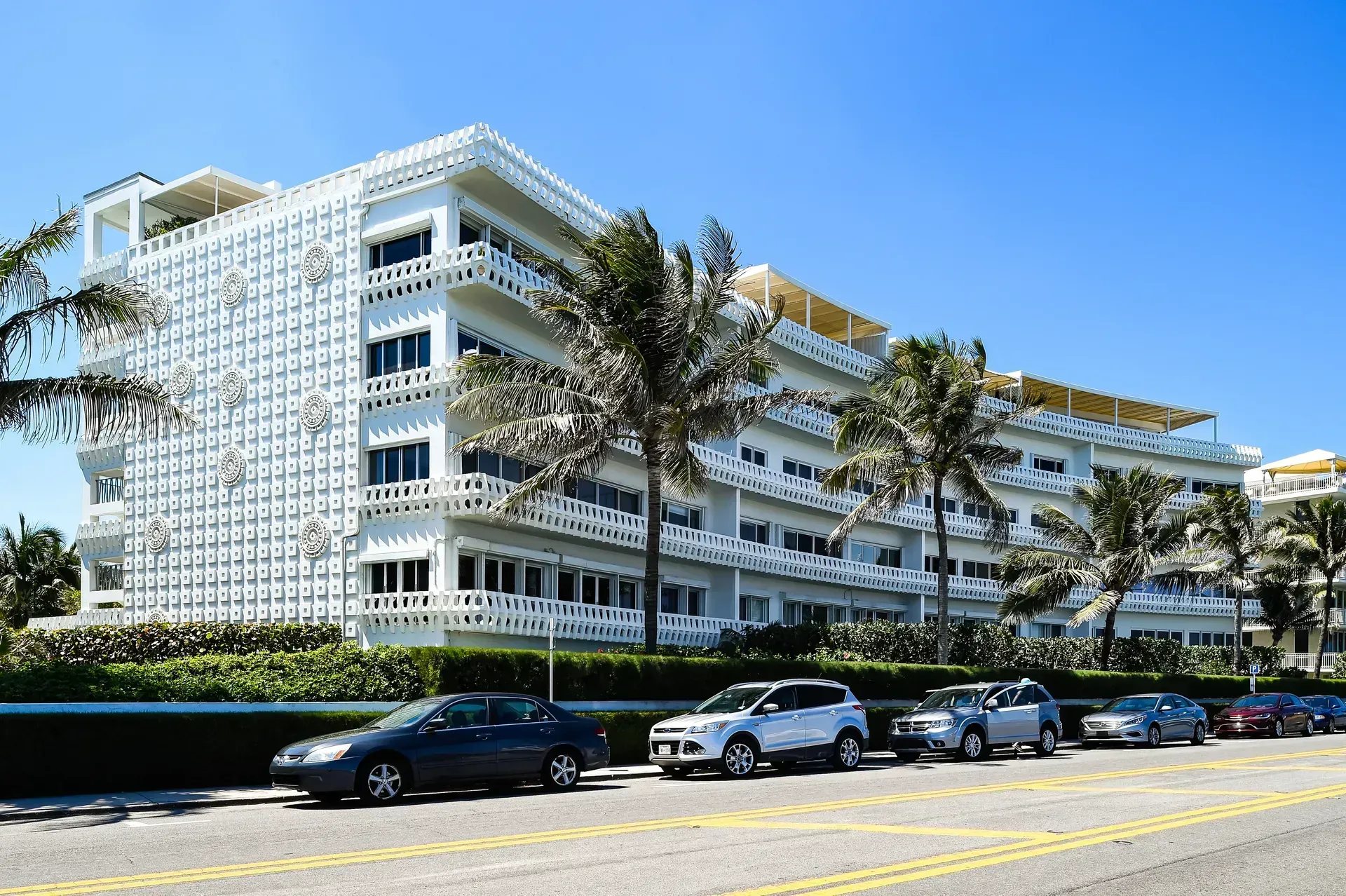 White multi-story hotel by the road with palm trees and parked cars under a clear blue sky