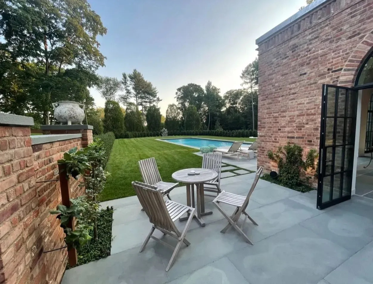 Patio with wooden chairs and table beside a brick house, overlooking a green lawn and garden.
