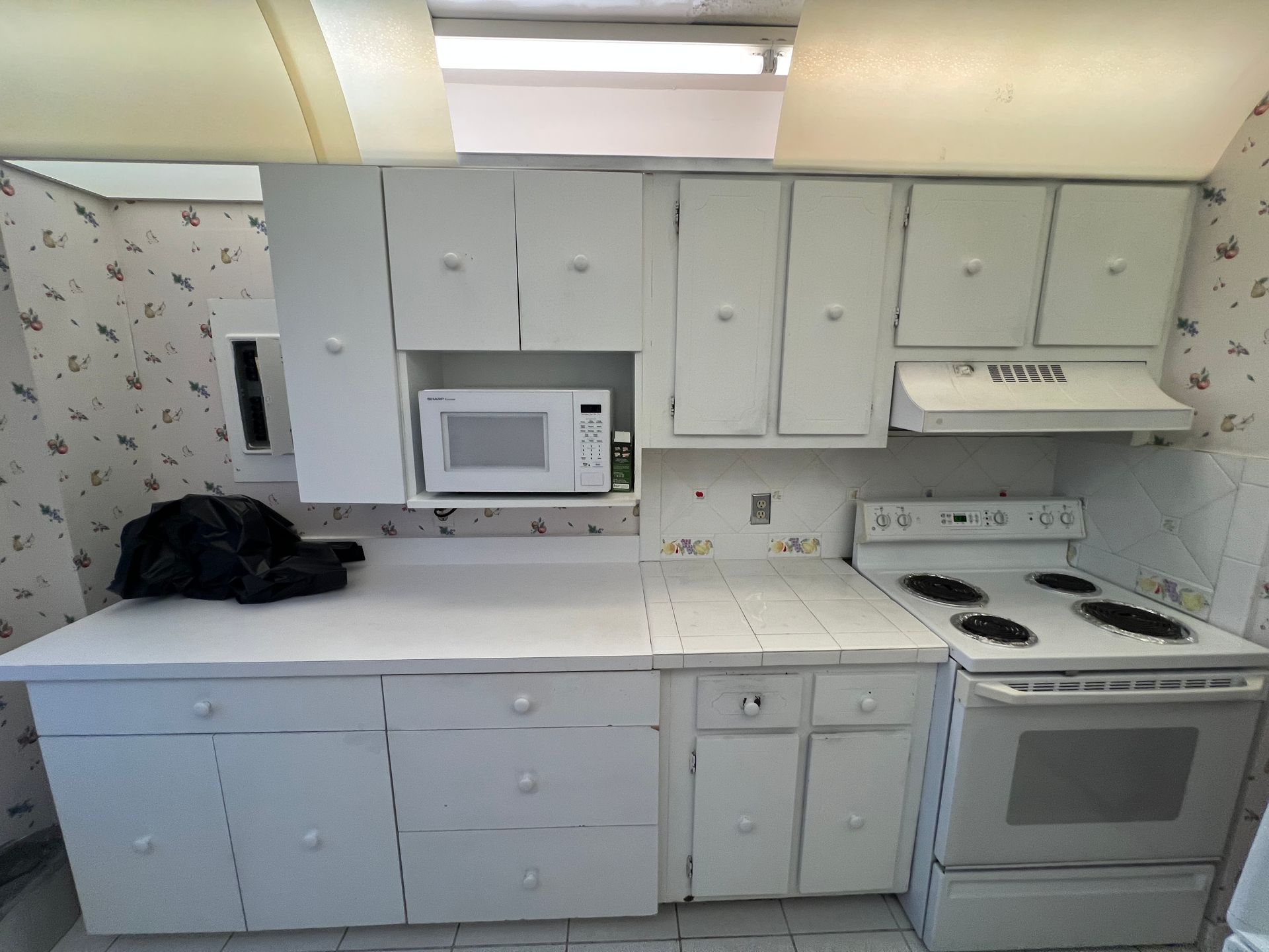 Small white kitchen with cabinets, microwave, stove, and a black bag on the counter.