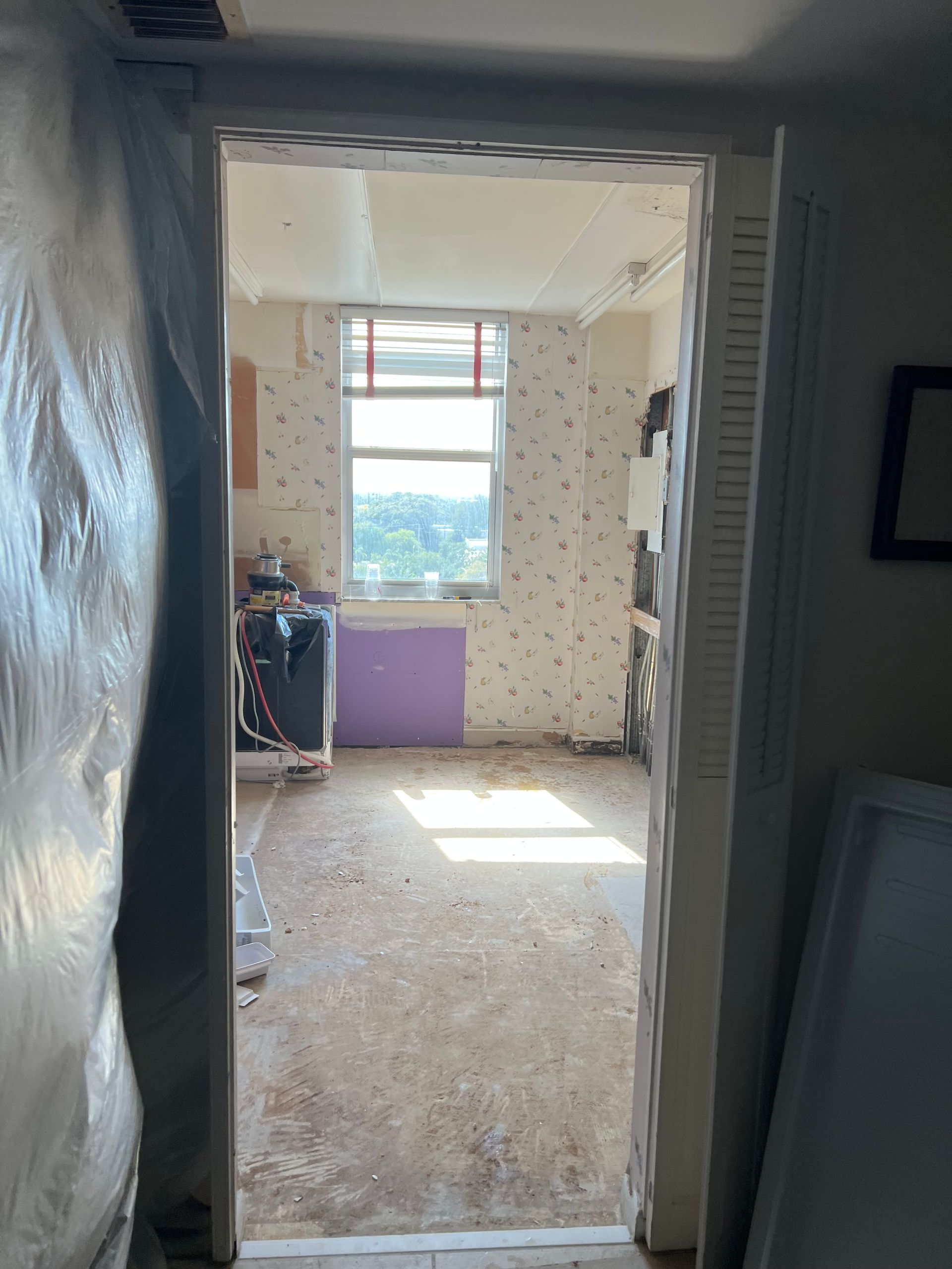 Dim room doorway with plastic sheeting, leading to a bright, partially renovated room with debris on the floor.