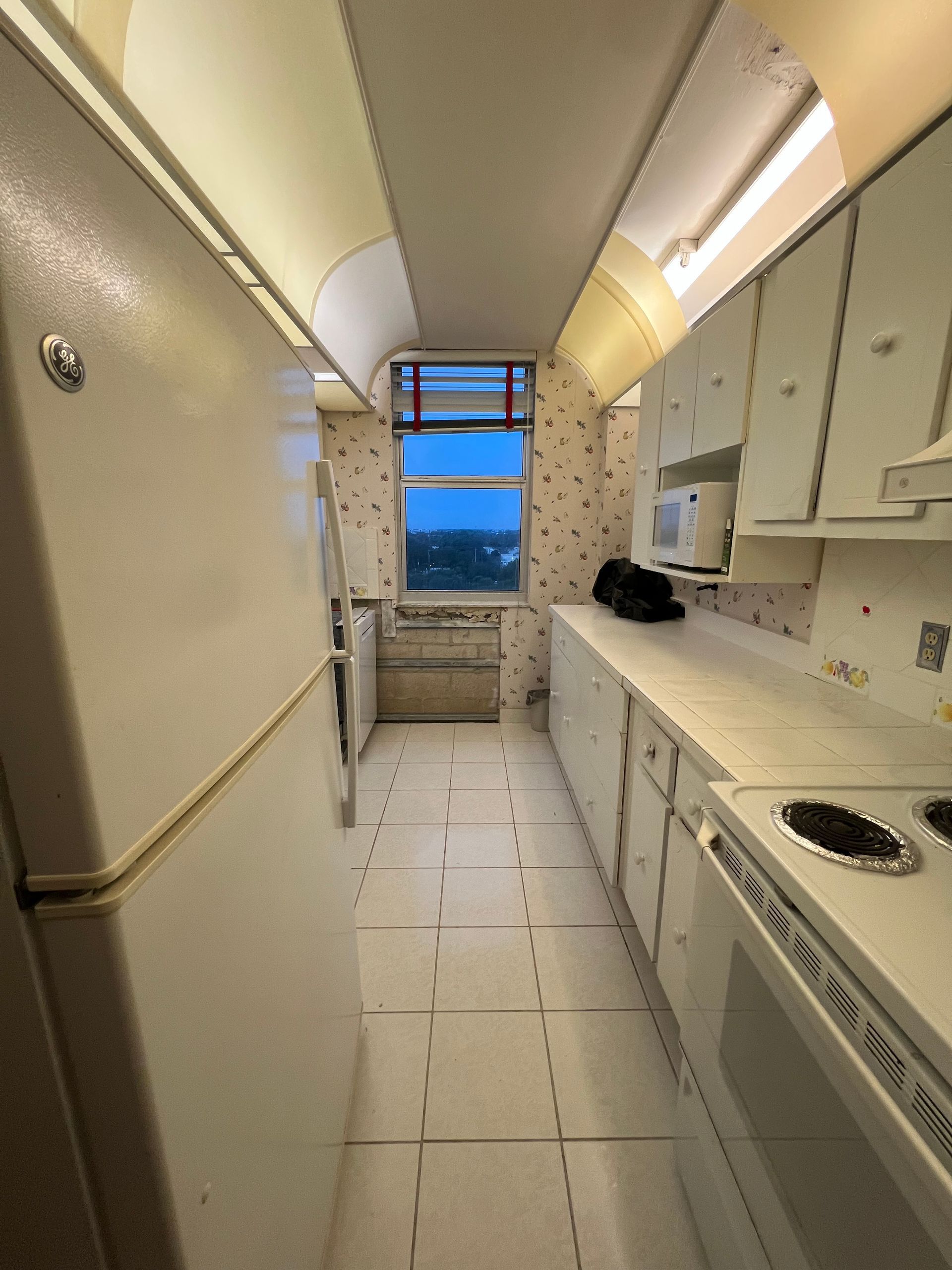Narrow galley kitchen with white cabinets, beige walls, tiled floor, and a window showing the ocean at dusk.