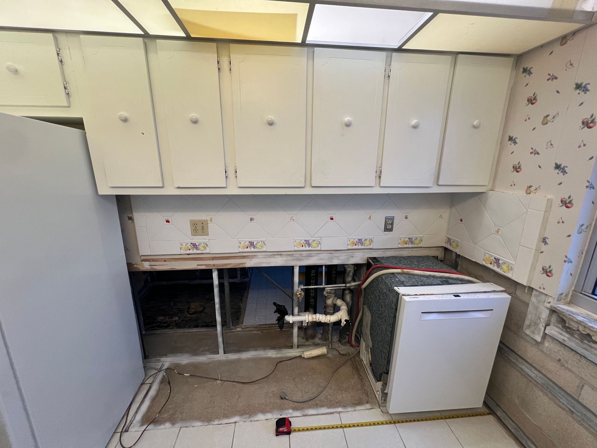 Damaged kitchen with missing lower cabinets, exposed plumbing, white upper cabinets, and a dishwasher on the right.