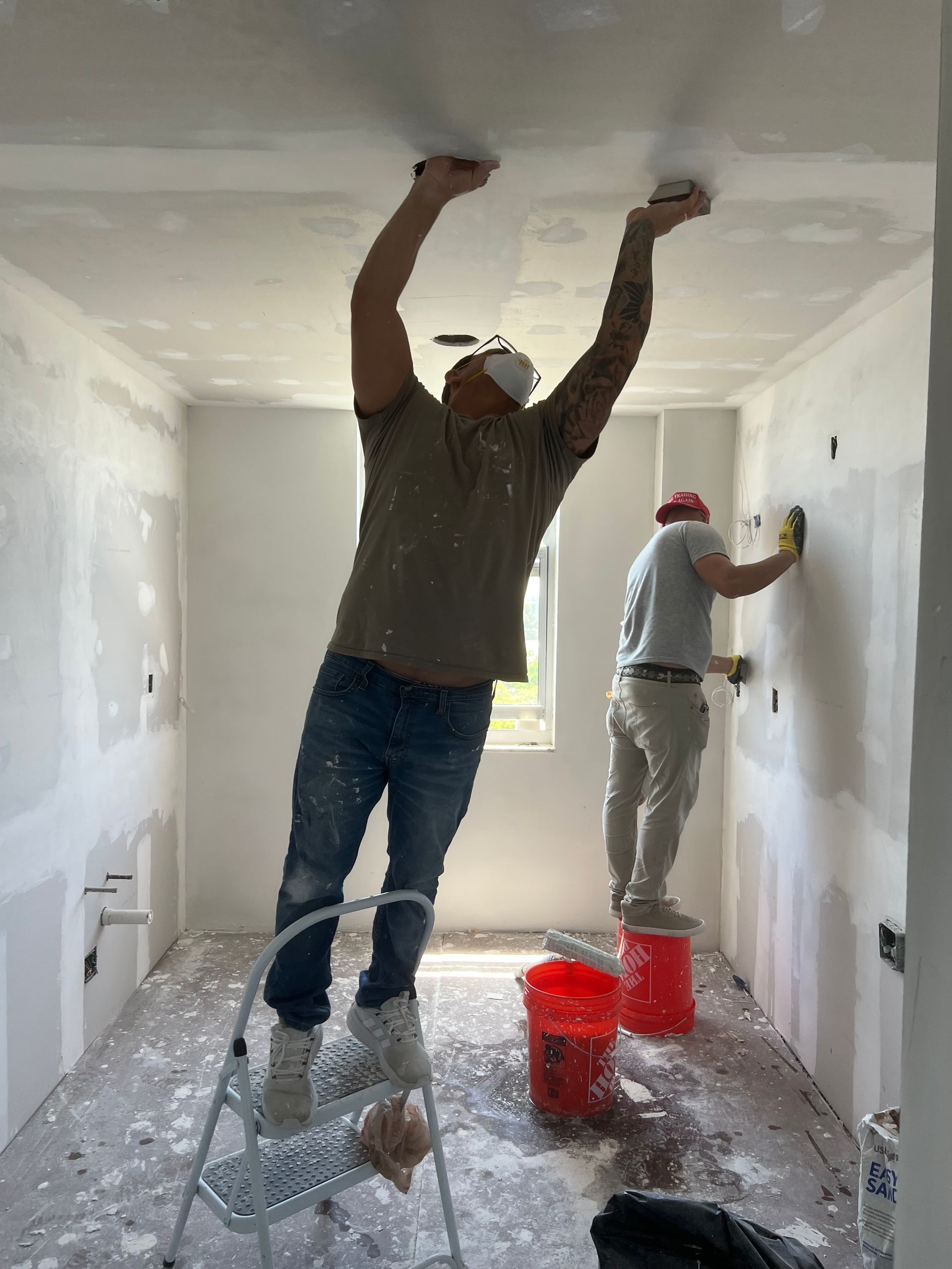 Two workers painting a room, one rolling the ceiling and one painting a wall near a window.