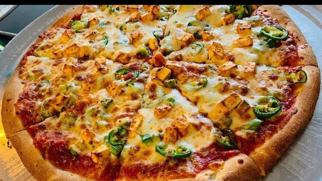 A round pizza with melted cheese, cubed paneer, and sliced jalapeños on a metal tray.