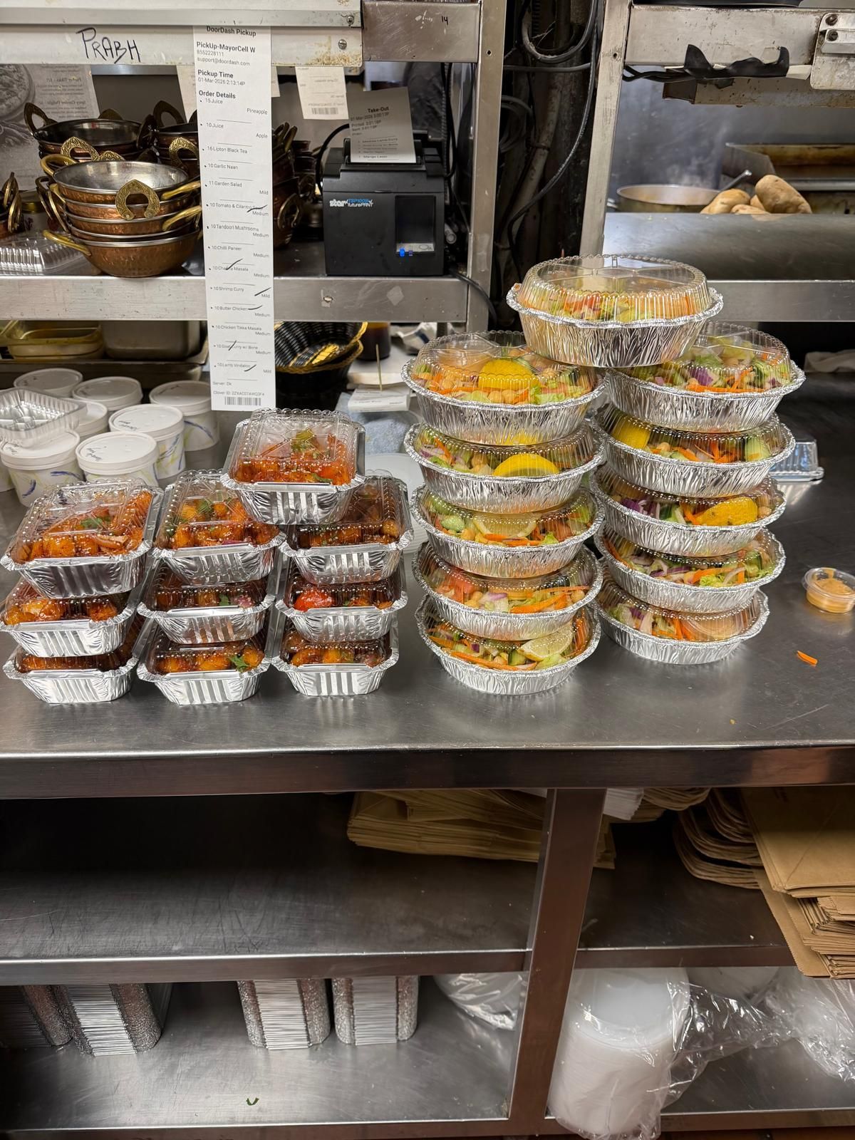 Many small foil takeout containers of various foods stacked on a stainless-steel commercial kitchen counter.