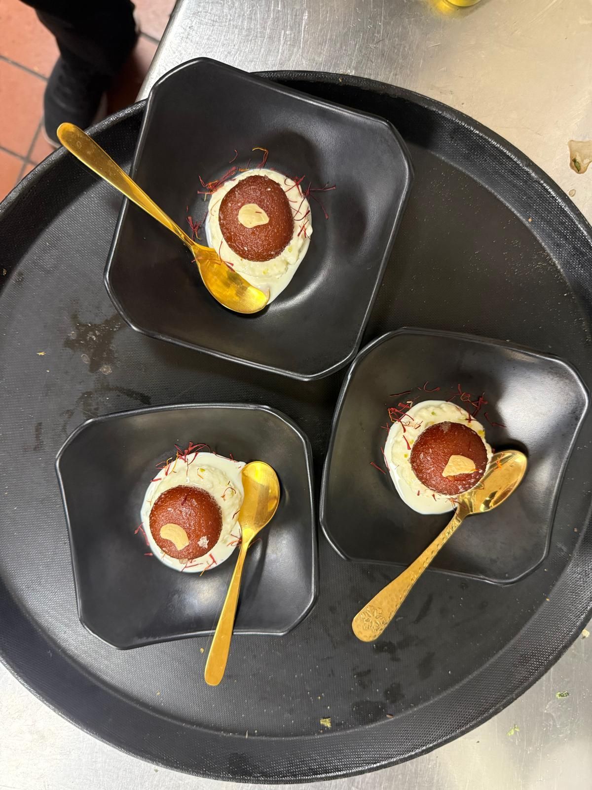 Three black bowls, each containing a single gulab jamun on a scoop of vanilla ice cream, sit on a tray with gold spoons.