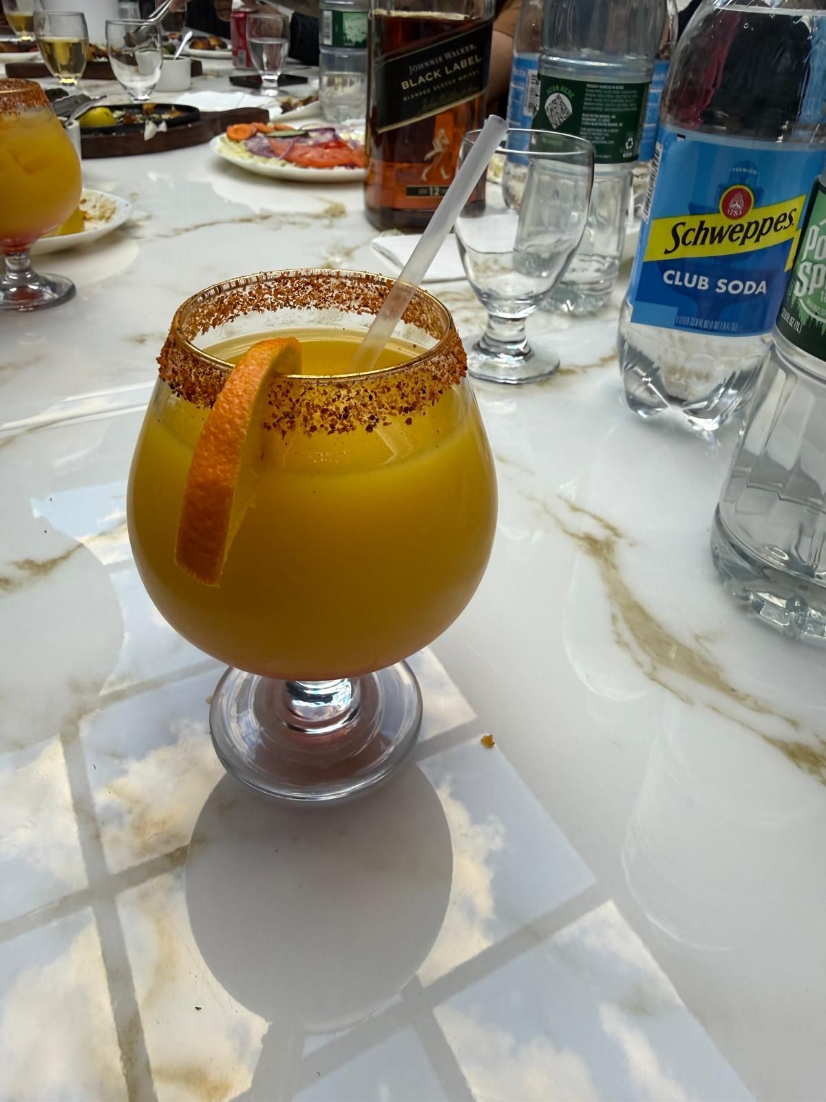 A bright orange cocktail in a wide glass with a chili-salt rim and orange slice, sitting on a marble table with drinks.