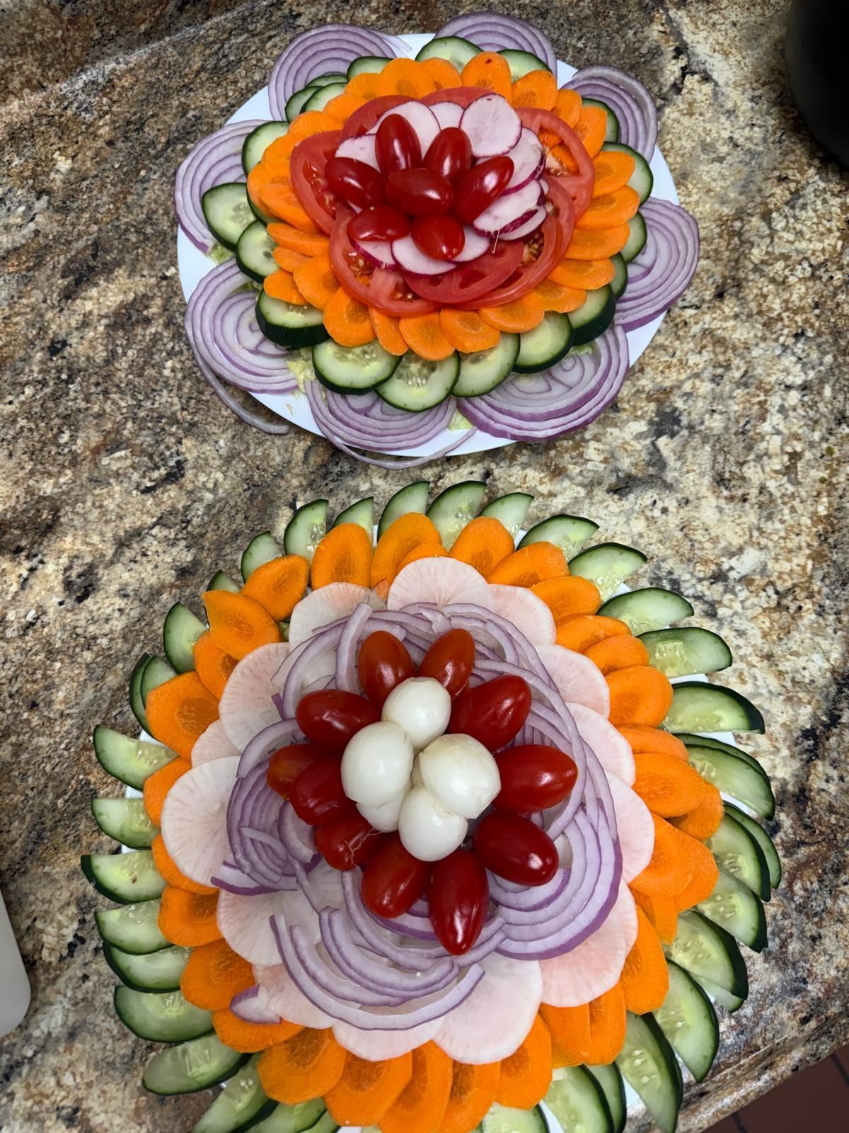 Two circular platters arranged with concentric, colorful layers of sliced vegetables and round deli meat on a stone counter.