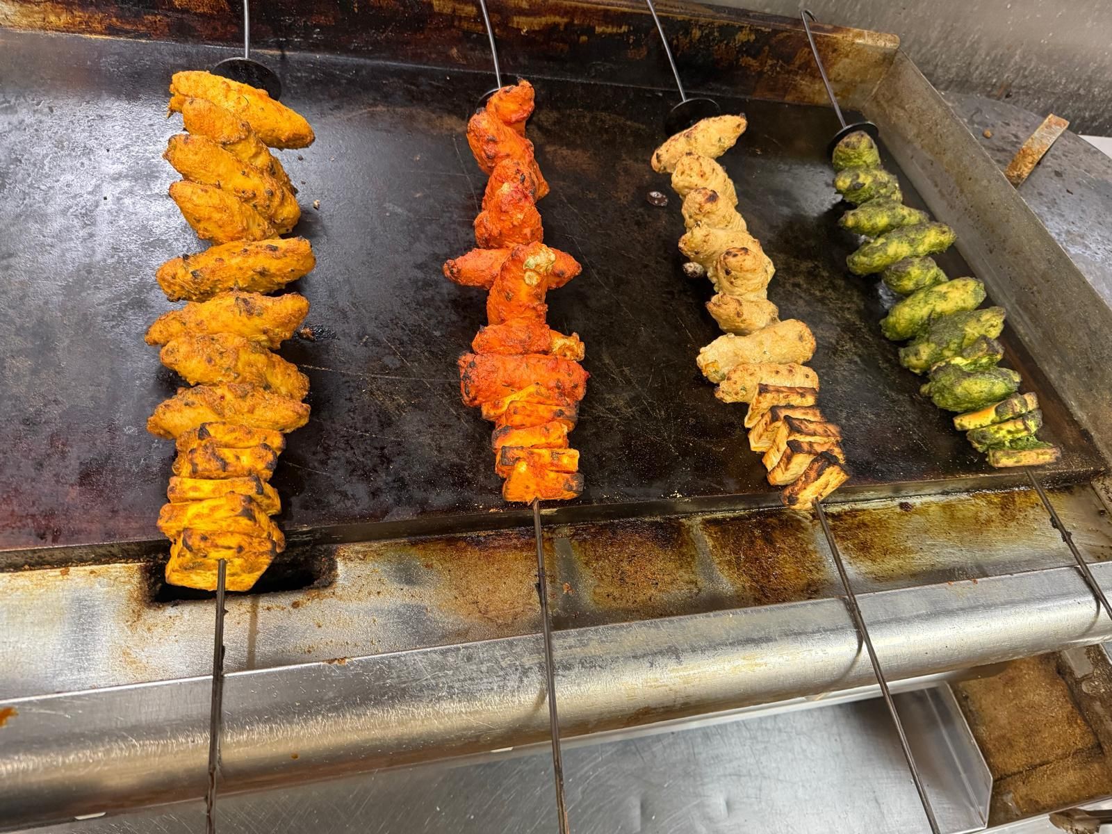 Four skewers of marinated meat and vegetables cooking on a flat-top grill, featuring yellow, red, tan, and green varieties.