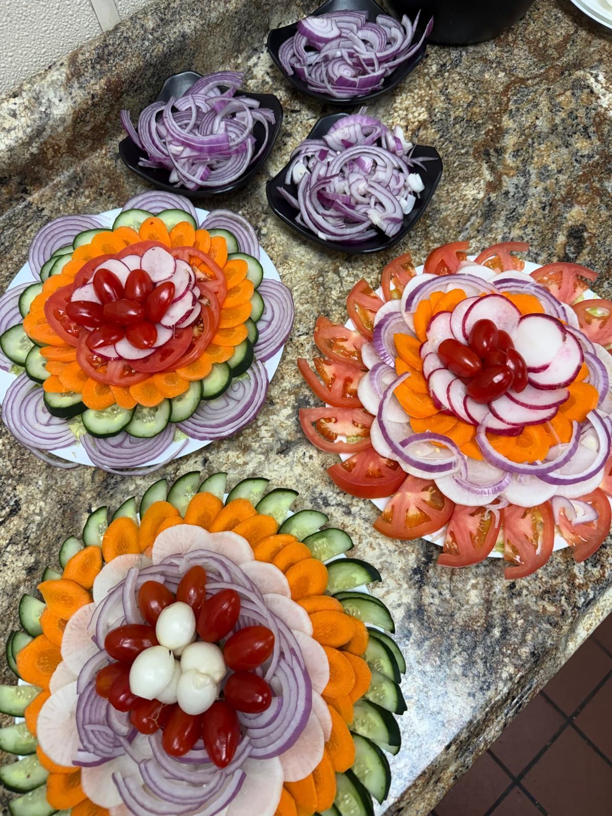 Three plates topped with circular, artistic arrangements of sliced cucumbers, carrots, tomatoes, radishes, and red onions.