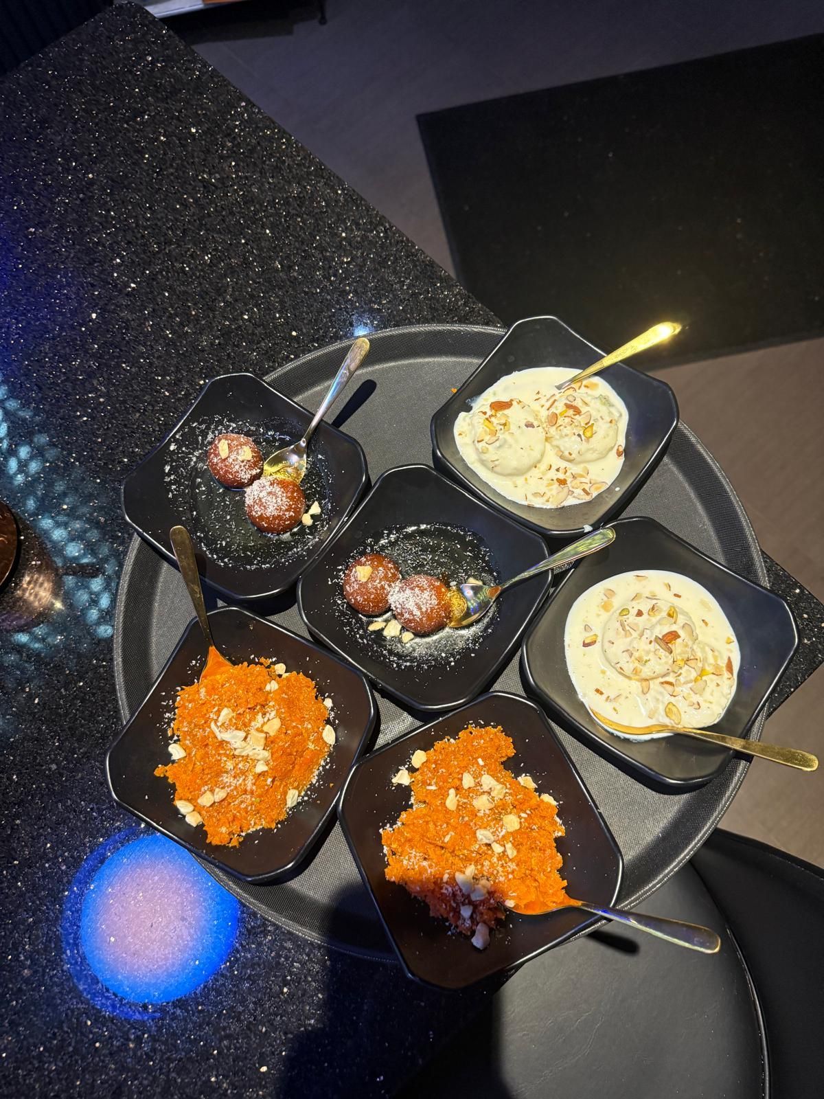 A tray containing five small, square bowls of various Indian desserts, including gajar halwa, ras malai, and gulab jamun.