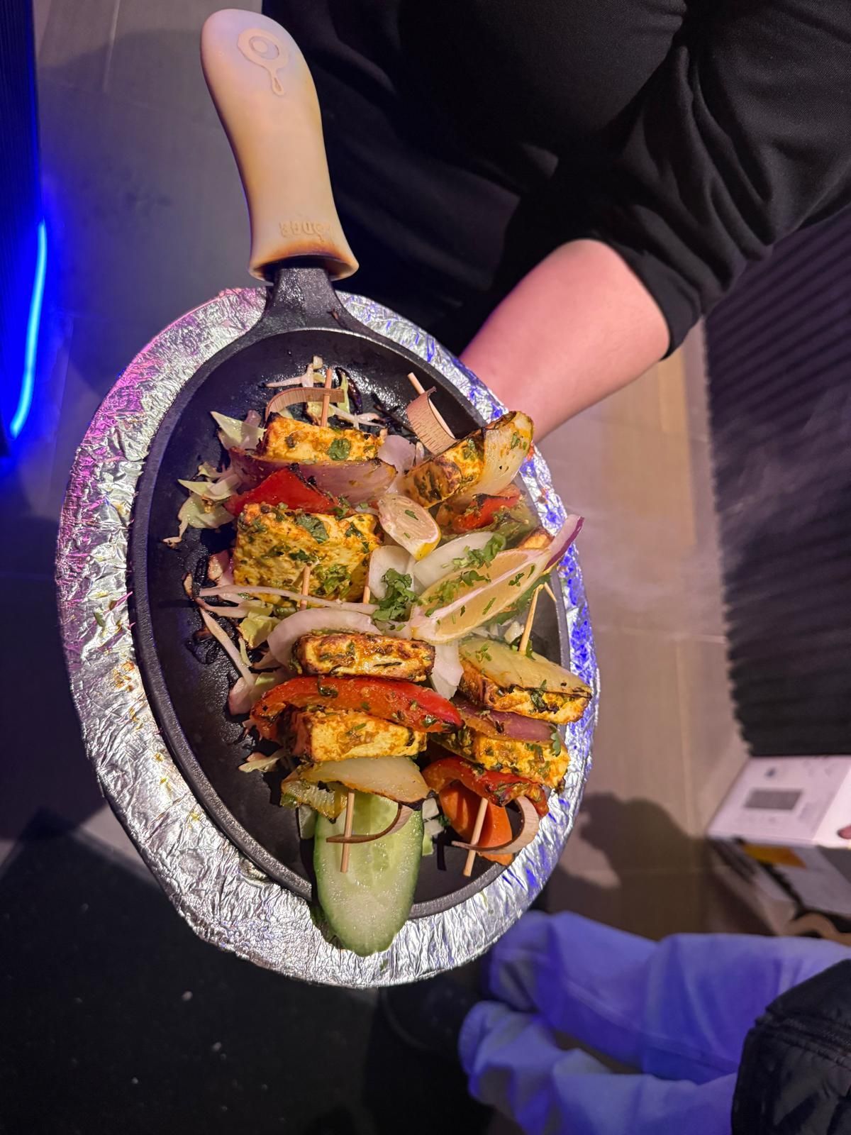 A hand holds a hot, oval cast-iron skillet featuring grilled paneer tikka skewers with peppers, onions, and cucumber slices.