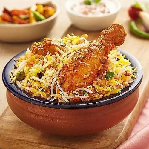 A clay pot filled with chicken biryani, topped with a chicken leg piece, served with side dishes on a wooden table.