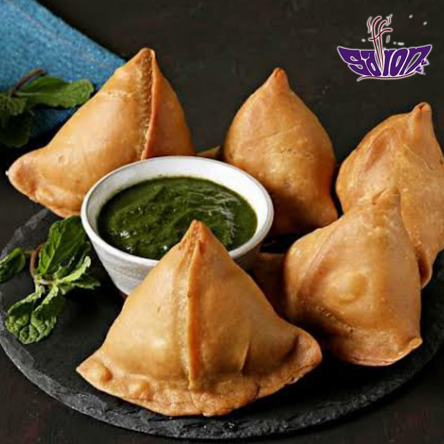 Five golden-brown samosas arranged around a small white bowl of green mint chutney on a dark, textured plate.