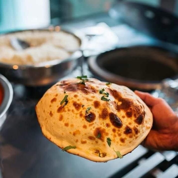 A hand holds a piece of golden-brown, puffed naan bread topped with fresh green herbs in a kitchen setting.