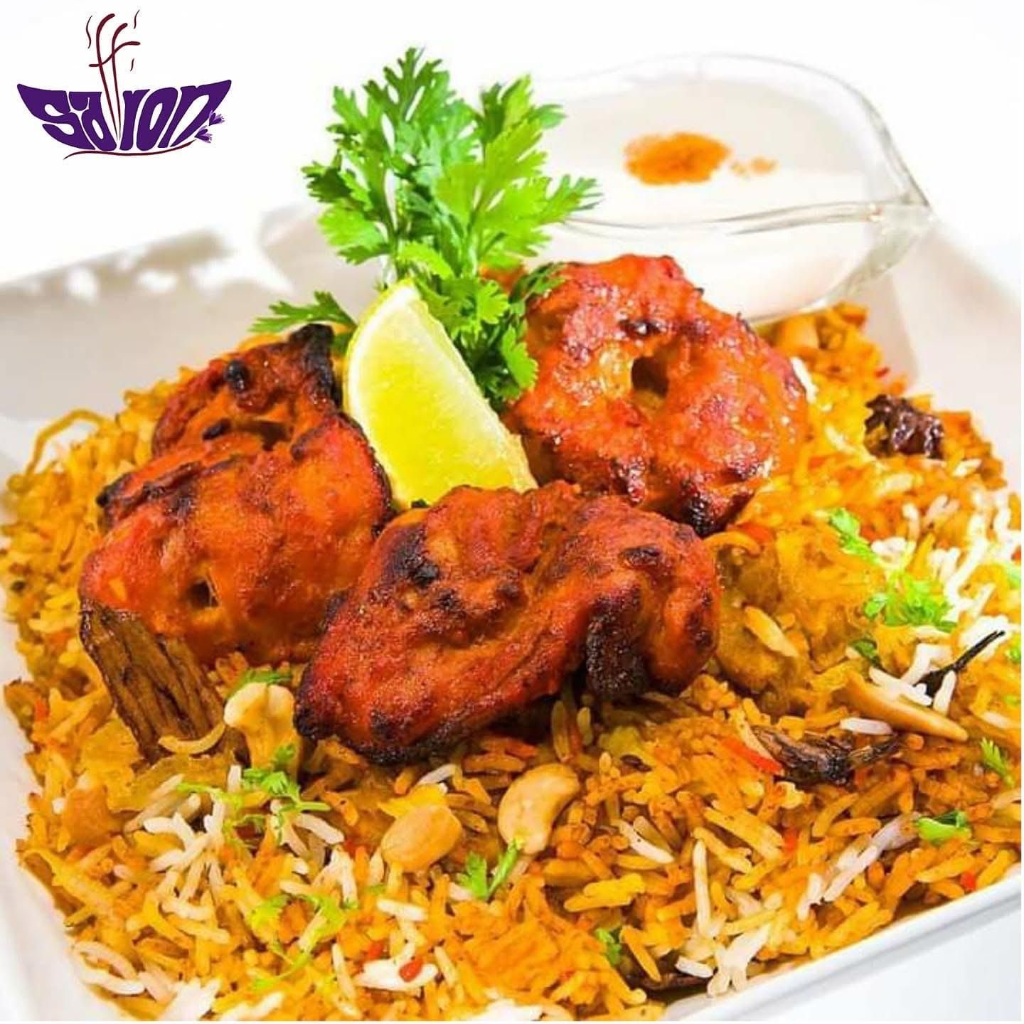 A serving of chicken tikka biryani topped with fresh cilantro, a lime wedge, and cashews, accompanied by a side of raita.