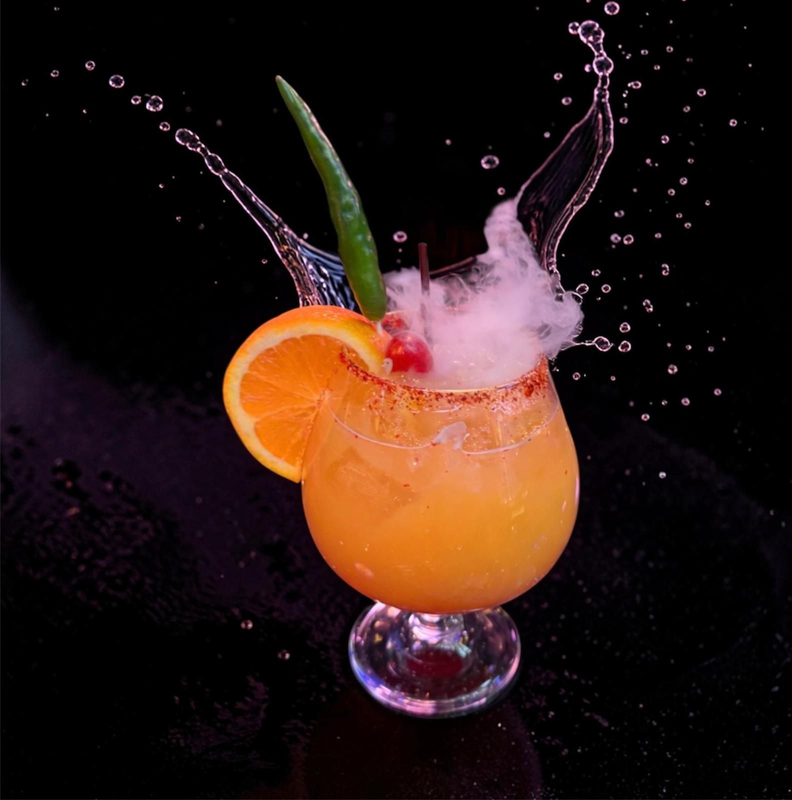 An orange cocktail in a stemmed glass with a chili pepper, orange slice, and a splash effect against a black background.