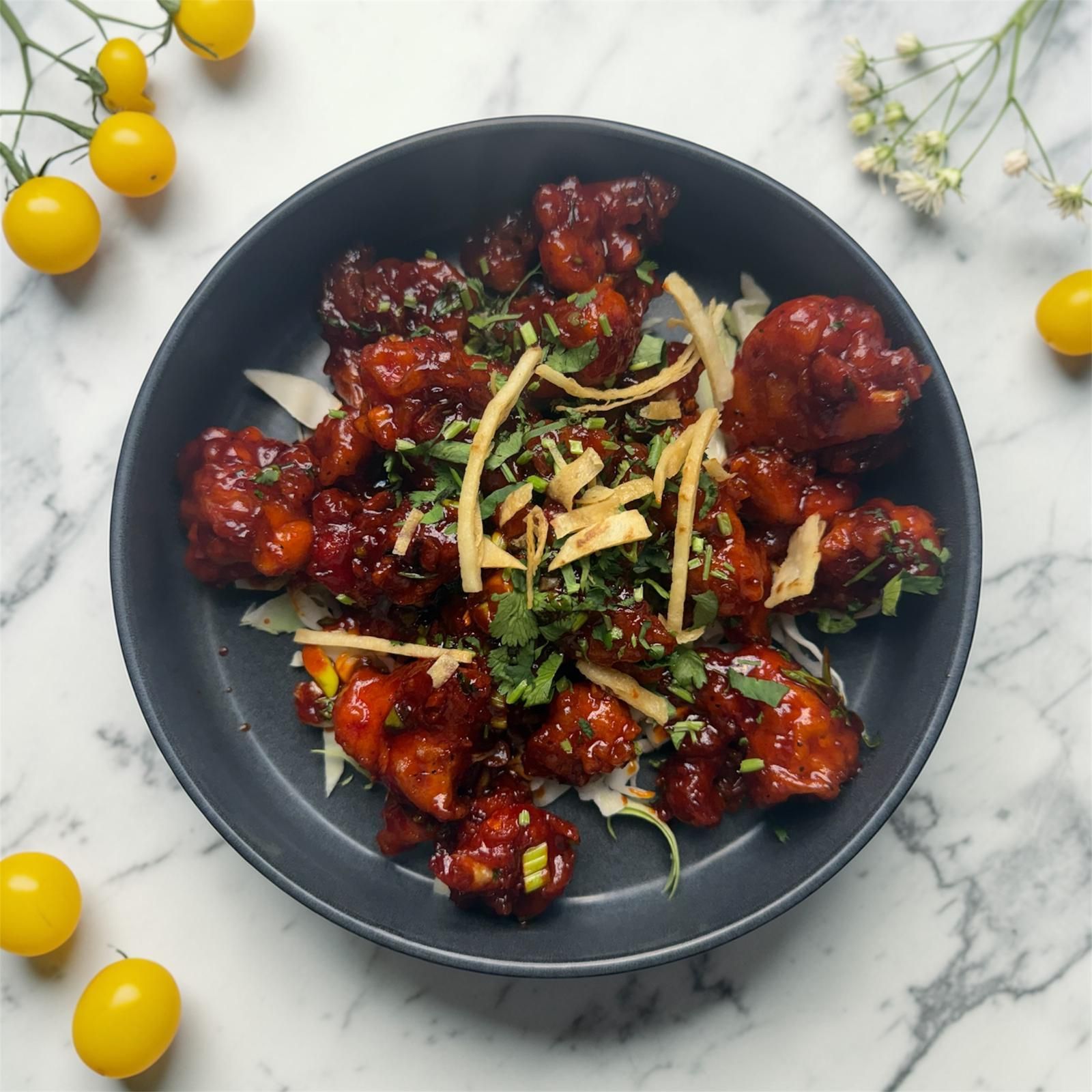 A bowl of crispy Gobi Manchurian topped with fresh herbs and ginger strips on a marble surface with yellow cherry tomatoes.