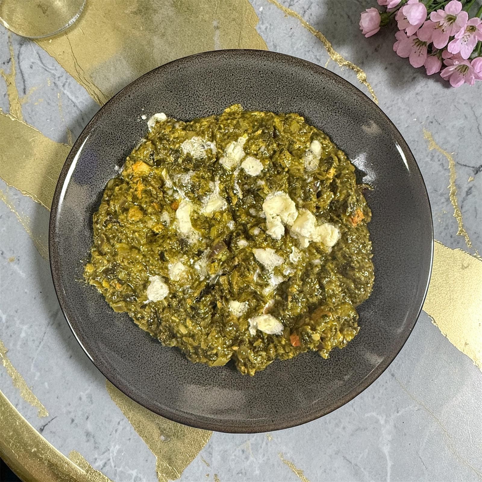 A plate of green vegetable stew or spinach risotto topped with crumbles of white cheese on a marble surface.