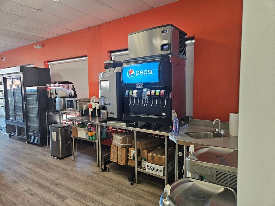 Pepsi fountain drink station in a cafeteria with shelves, coolers, and orange walls