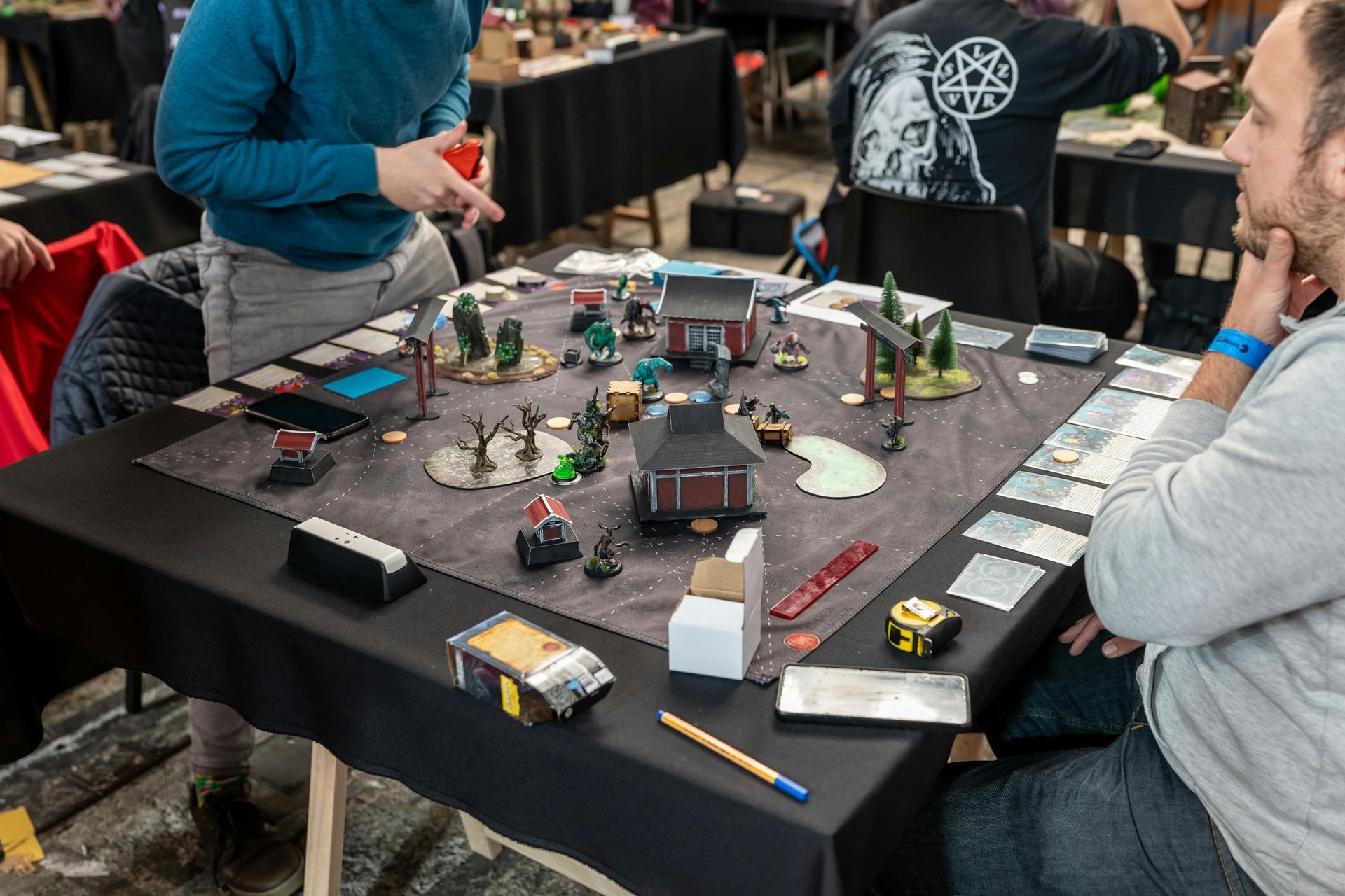 Tabletop game in progress on a black mat with miniatures and cards; two players sit around the table.