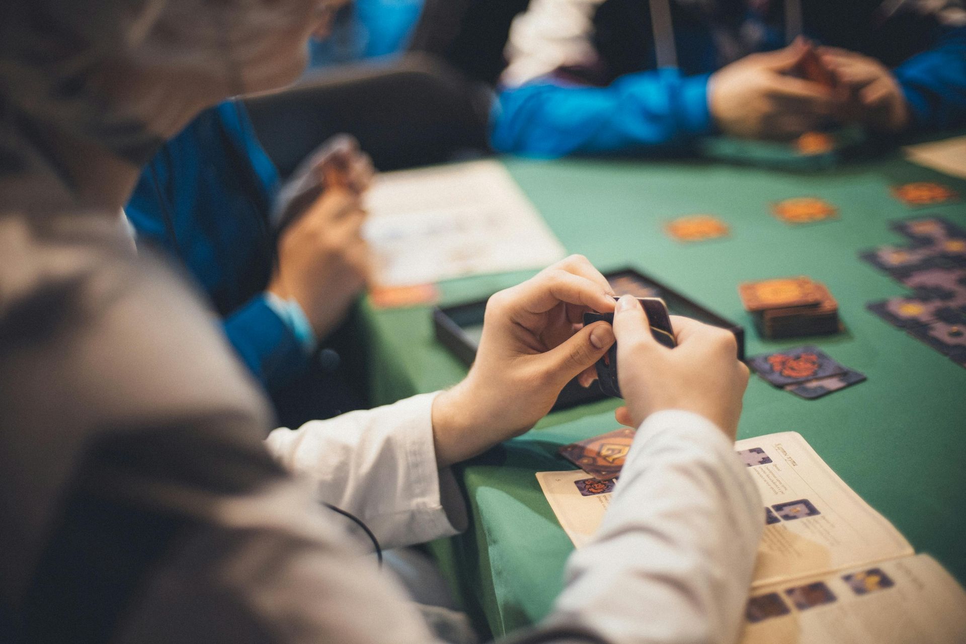 People playing a card game at a green table, with hands holding cards and game pieces spread out.