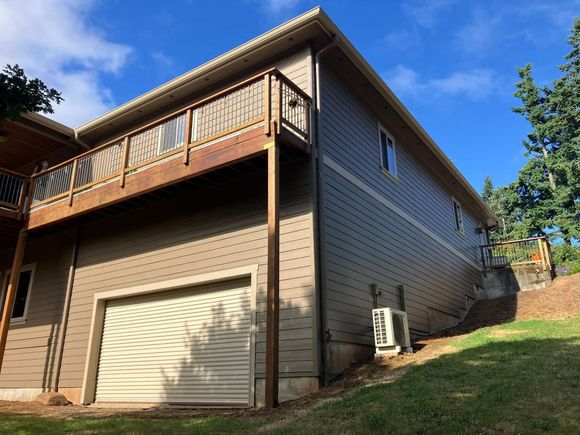 A two-story house with grey siding, a wooden deck, a garage door on the ground level, and an outdoor AC unit.