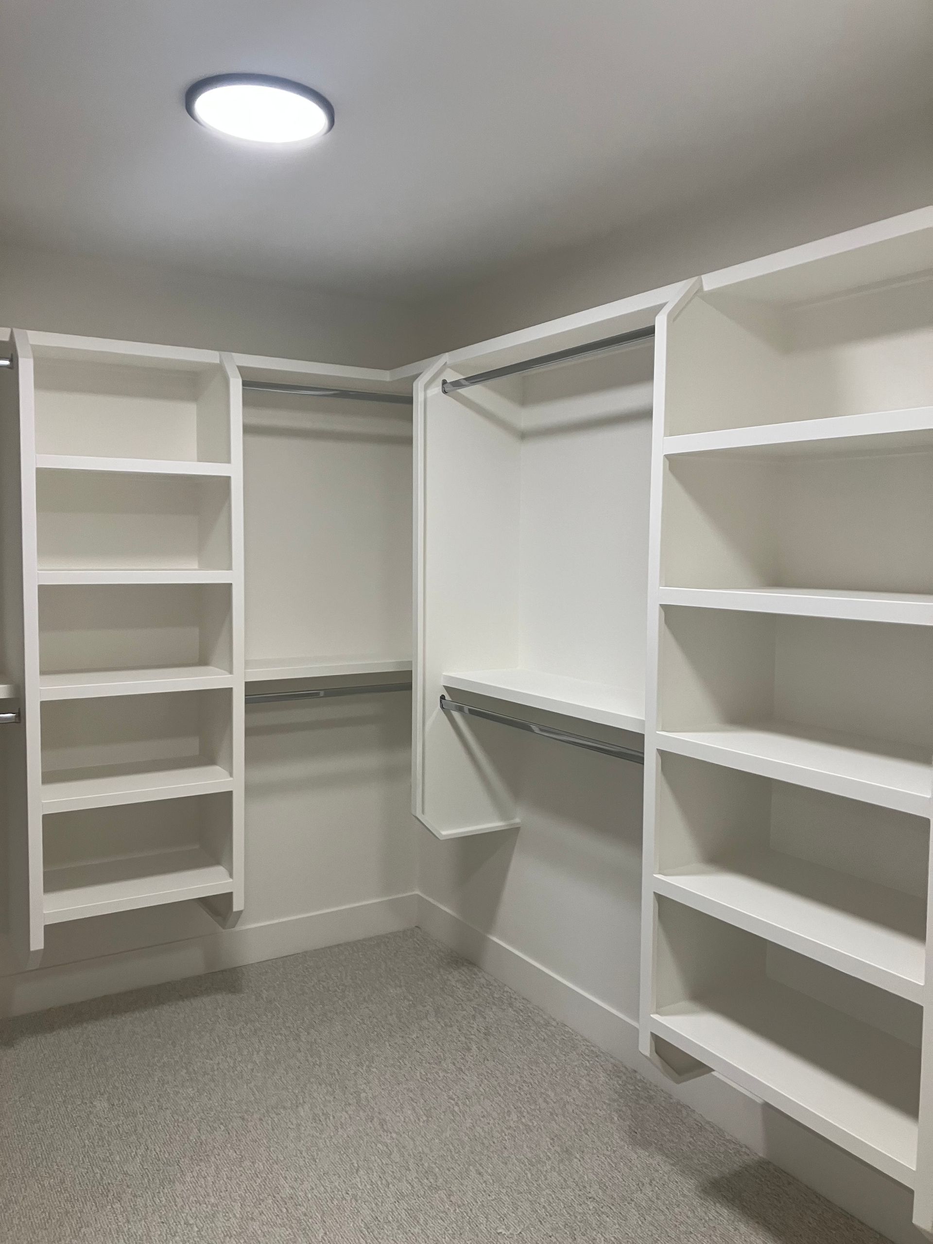 Empty walk-in closet with white built-in shelves, hanging rods, and gray carpet.