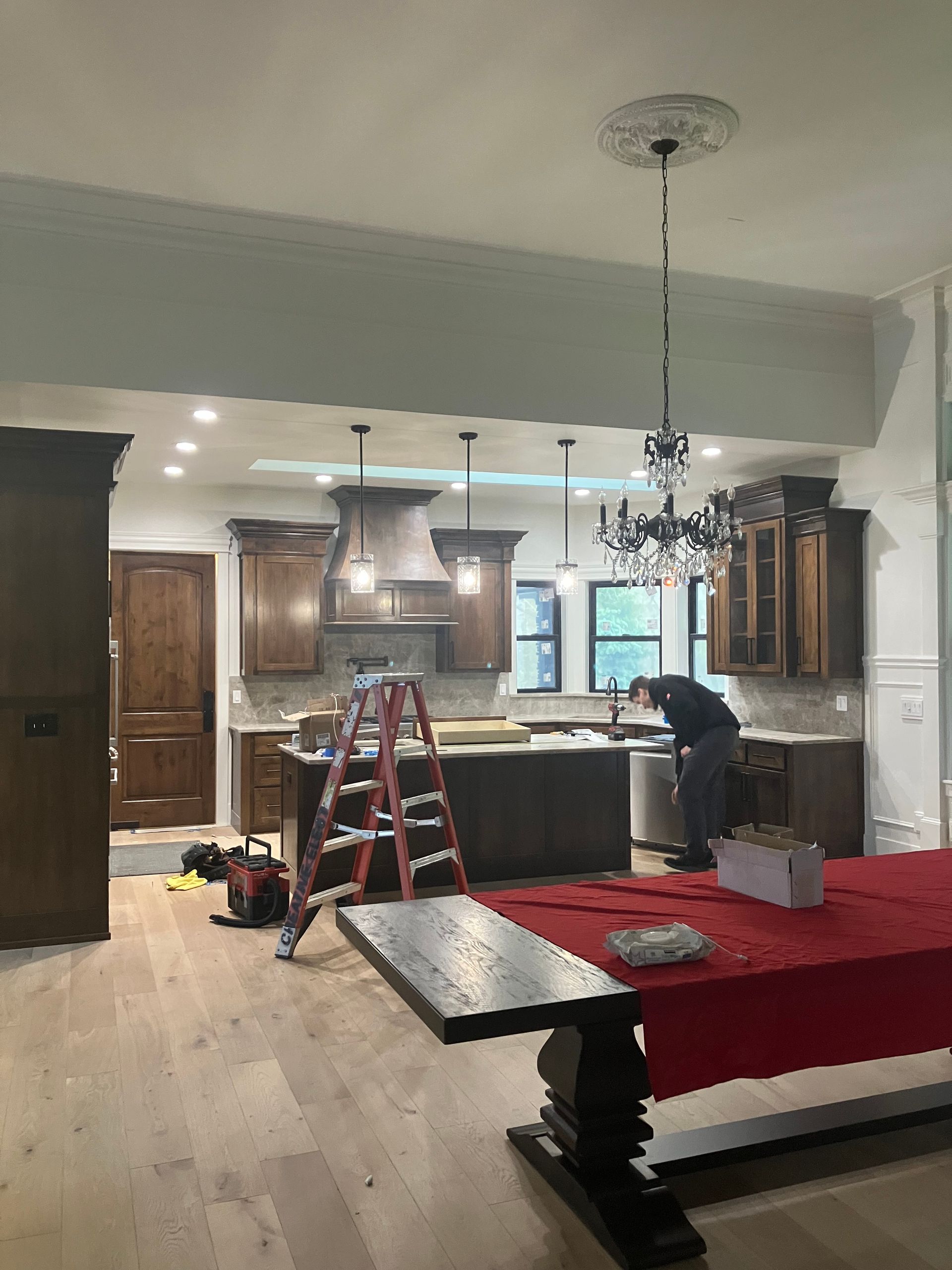 Kitchen under renovation with ladder, tools, and red-covered table beneath a chandelier