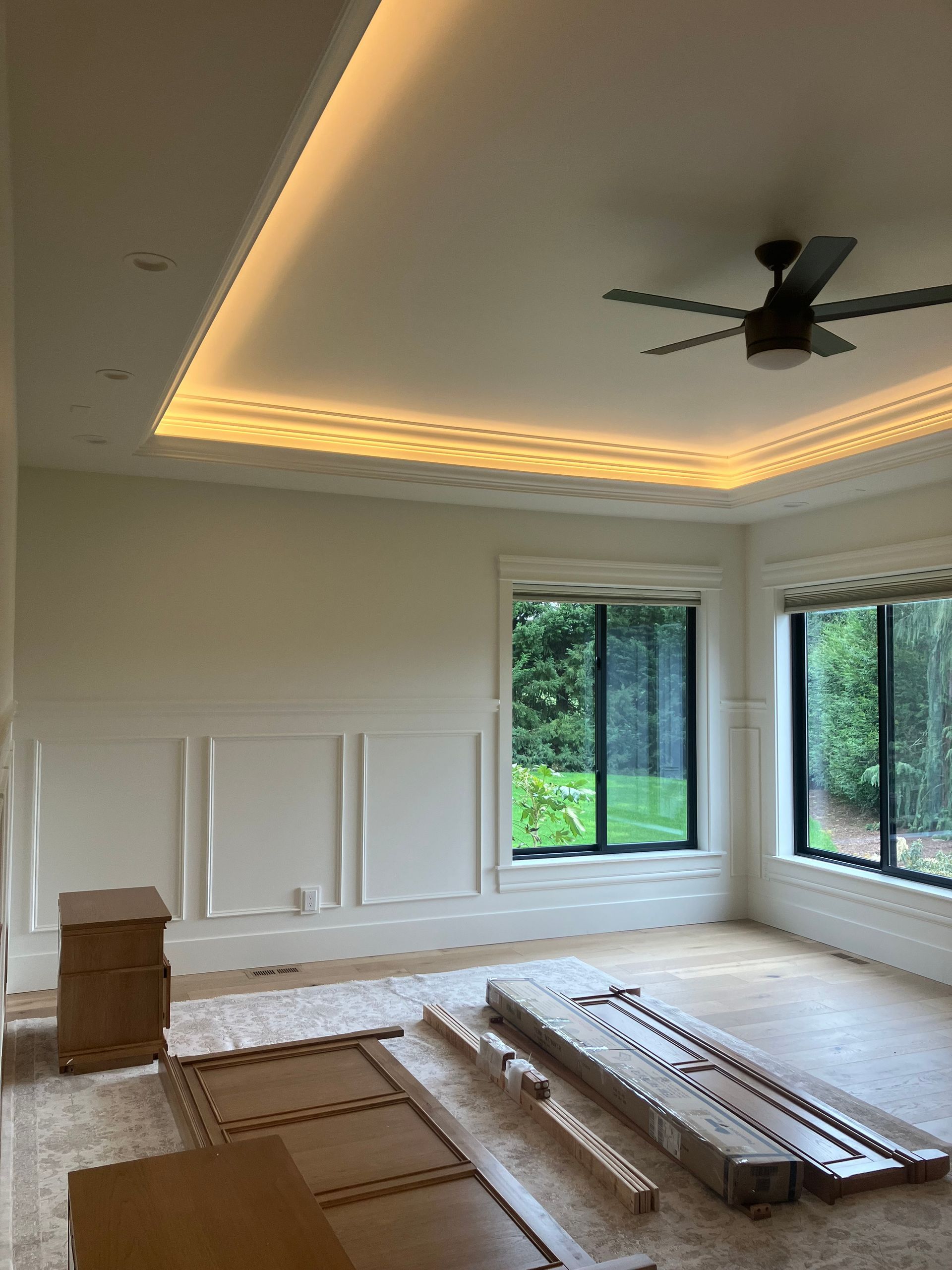 Empty living room with recessed ceiling lights, ceiling fan, and windows; furniture wrapped and stacked on floor