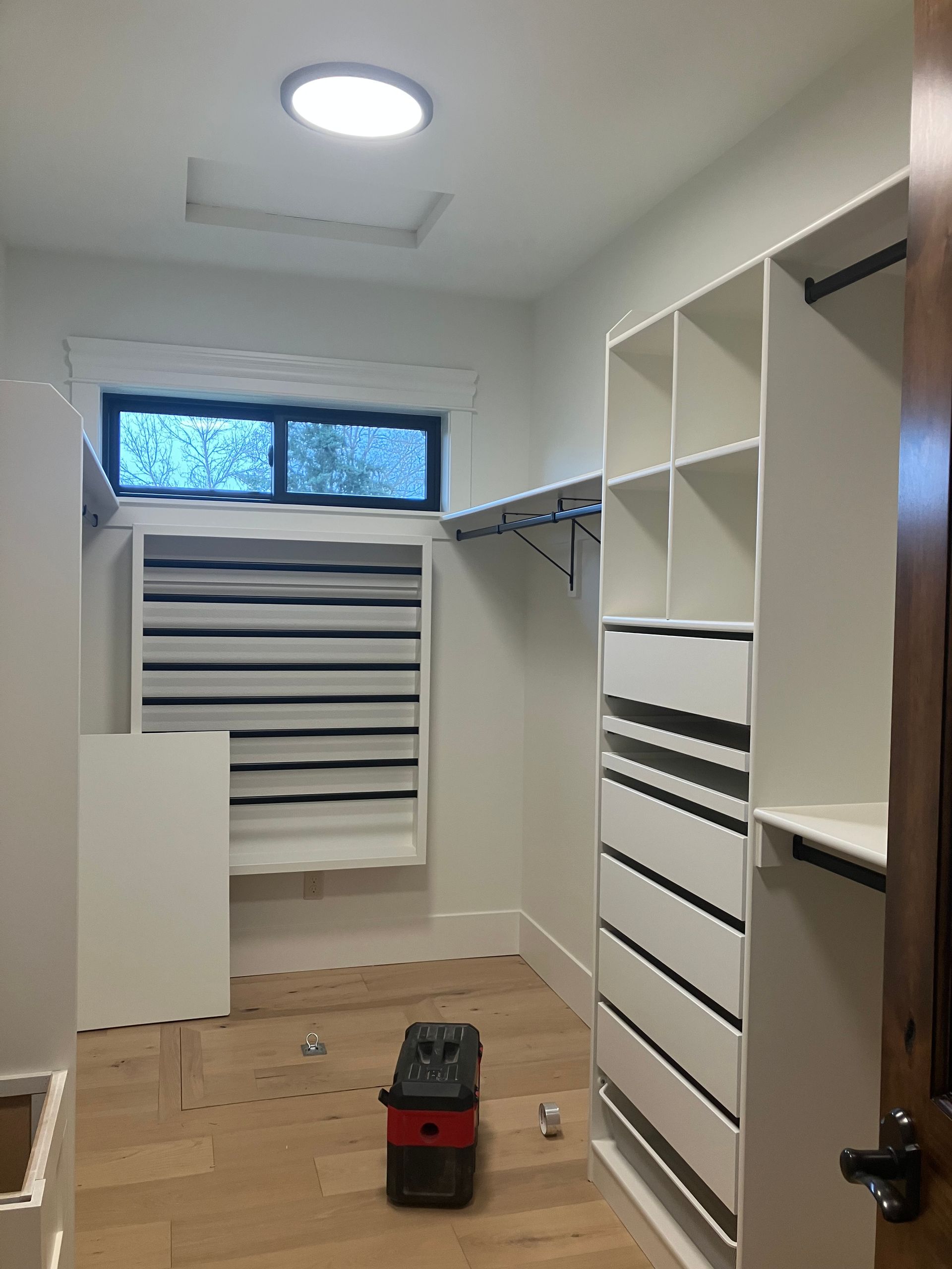 Walk-in closet under renovation with white shelving, drawers, and a striped window blind.