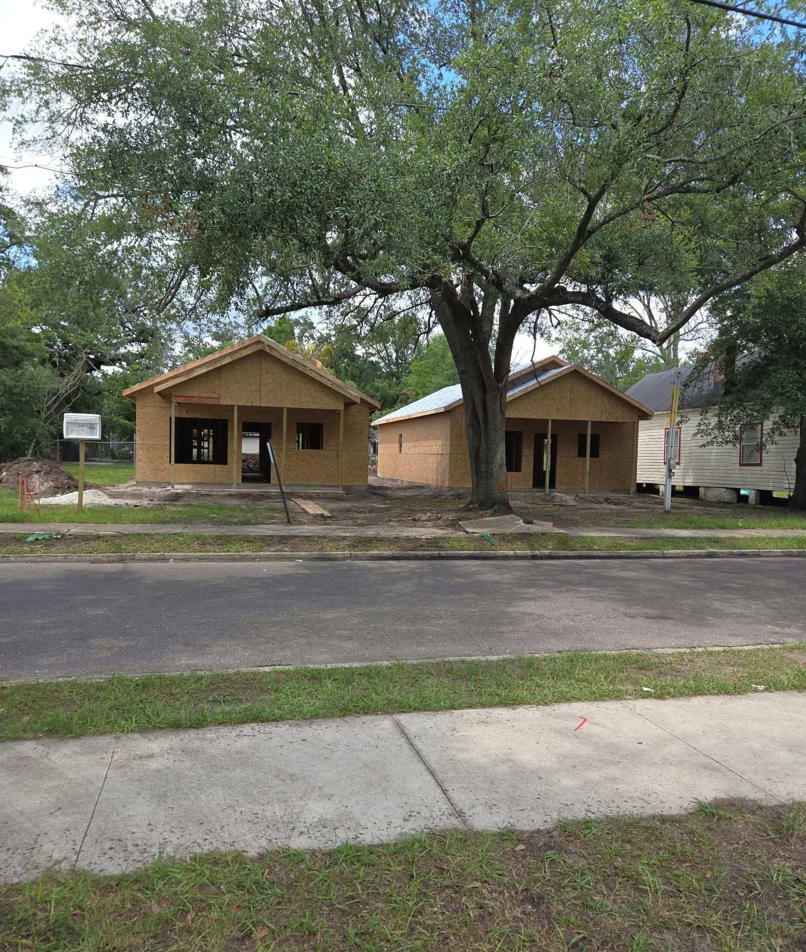 Two tan houses with front porches under trees, viewed across a road and sidewalk.