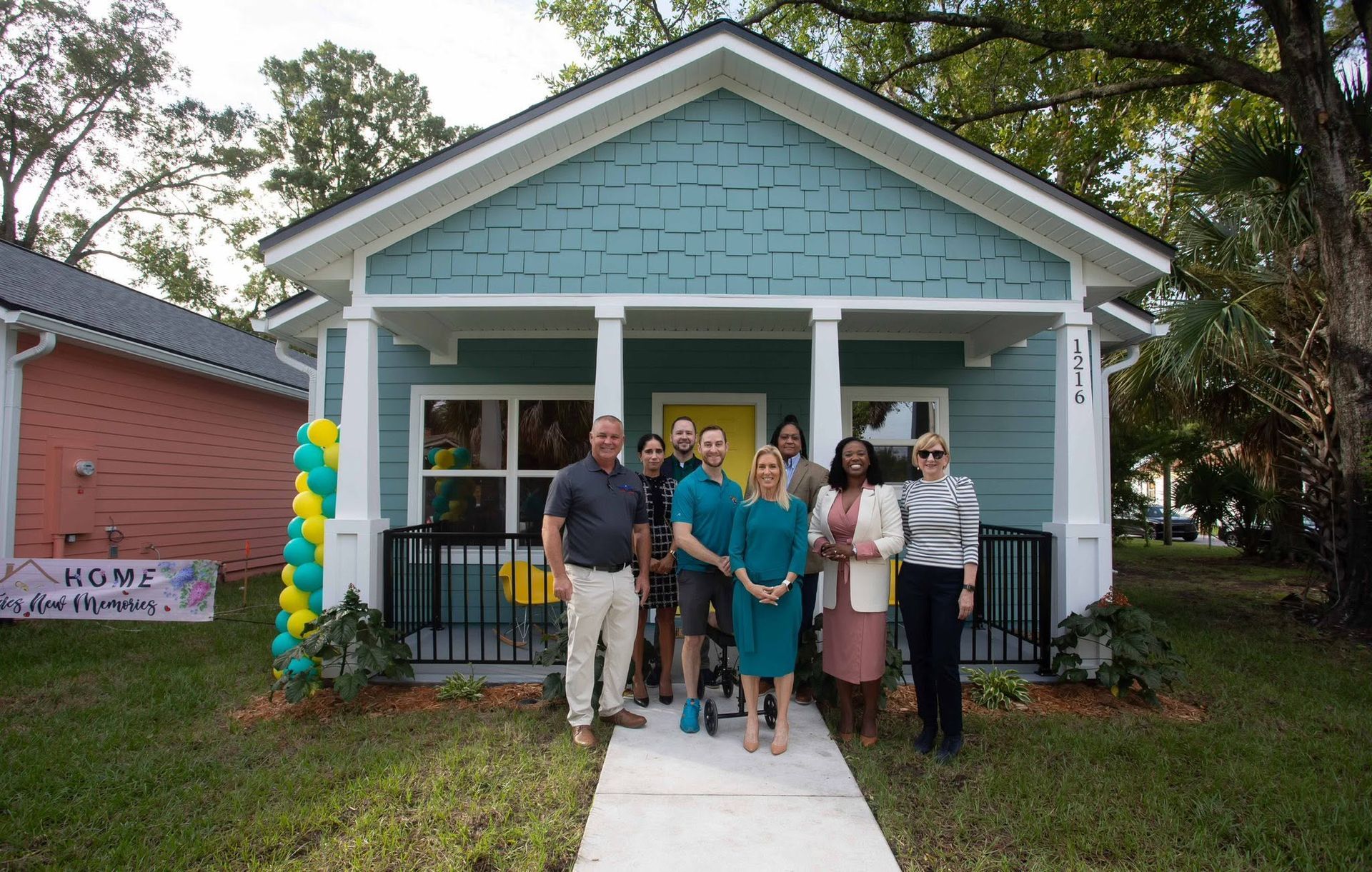 Group posing on a white walkway in front of a blue house with balloons and a ribbon-cutting setup