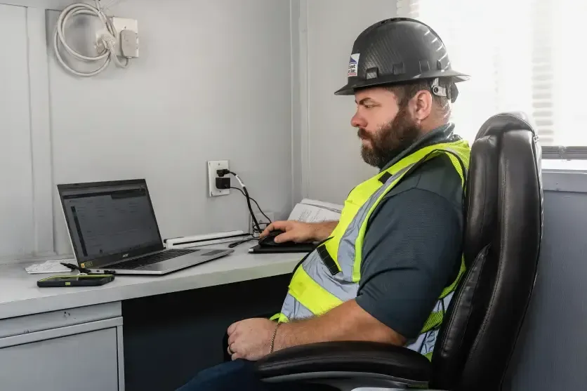 Man in hard hat and safety vest working at a desk with laptop in a small office-like room