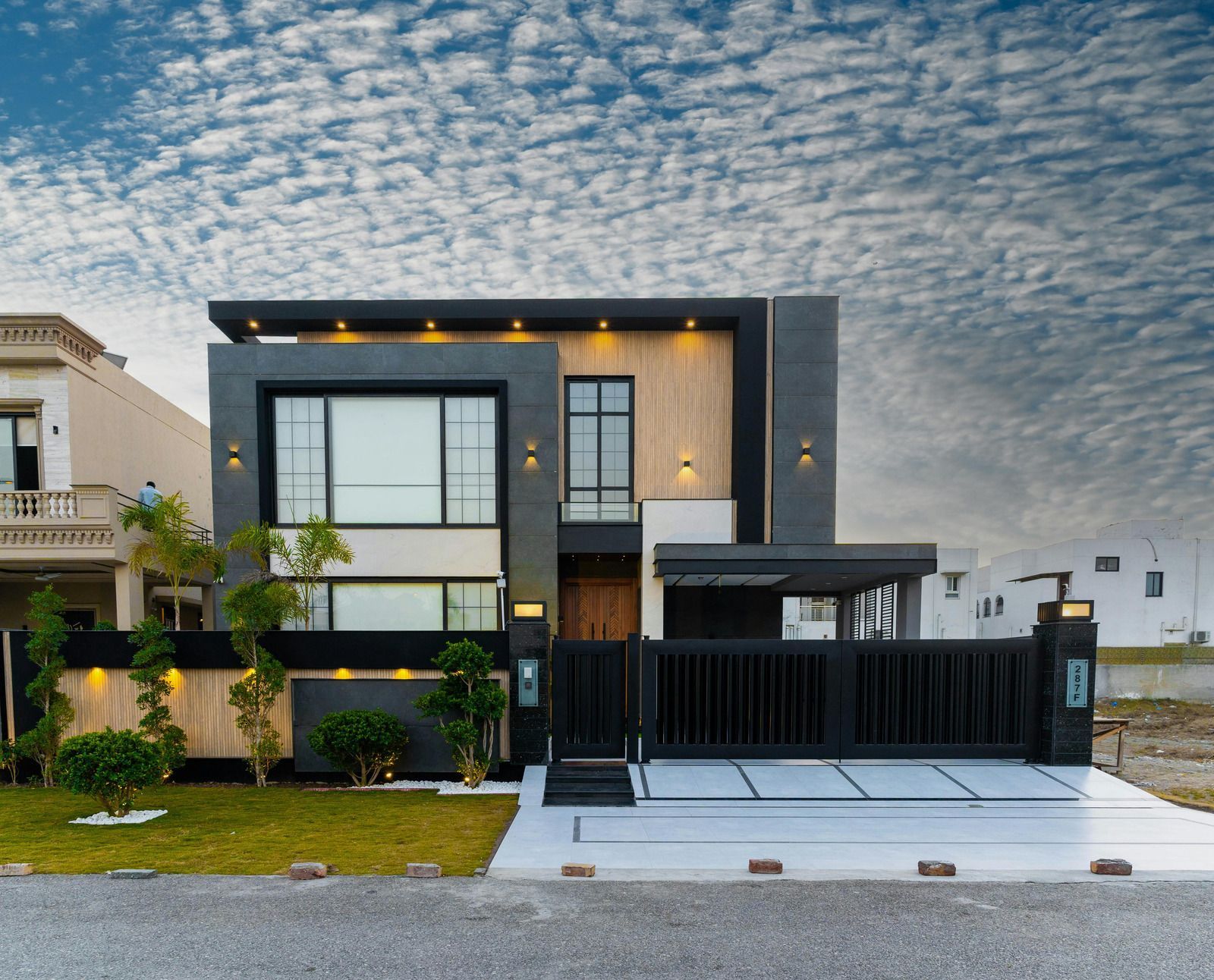Modern two-story house with dark facade, lit windows, front lawn, and black gate under a cloudy sky