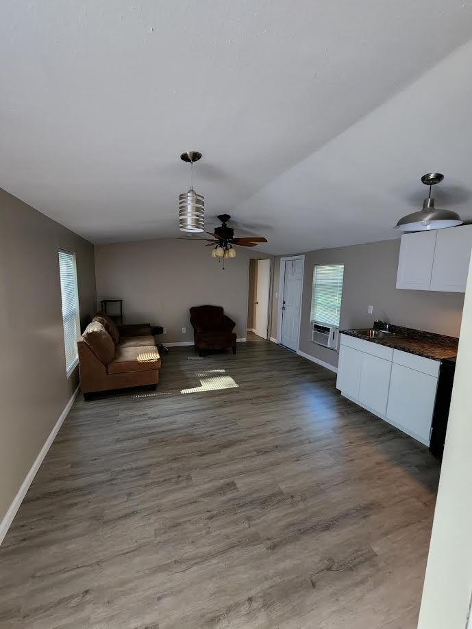 Empty living room with wood floors, brown sofa, recliner, ceiling fans, and a small kitchen area.