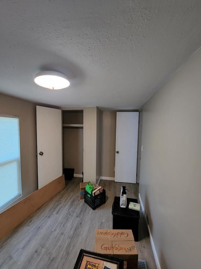 Small empty room with beige walls, gray floor, ceiling light, and boxes near the back wall.