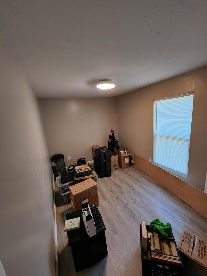 Empty room with boxes and furniture along the walls, light wood floor, and a window with blinds.